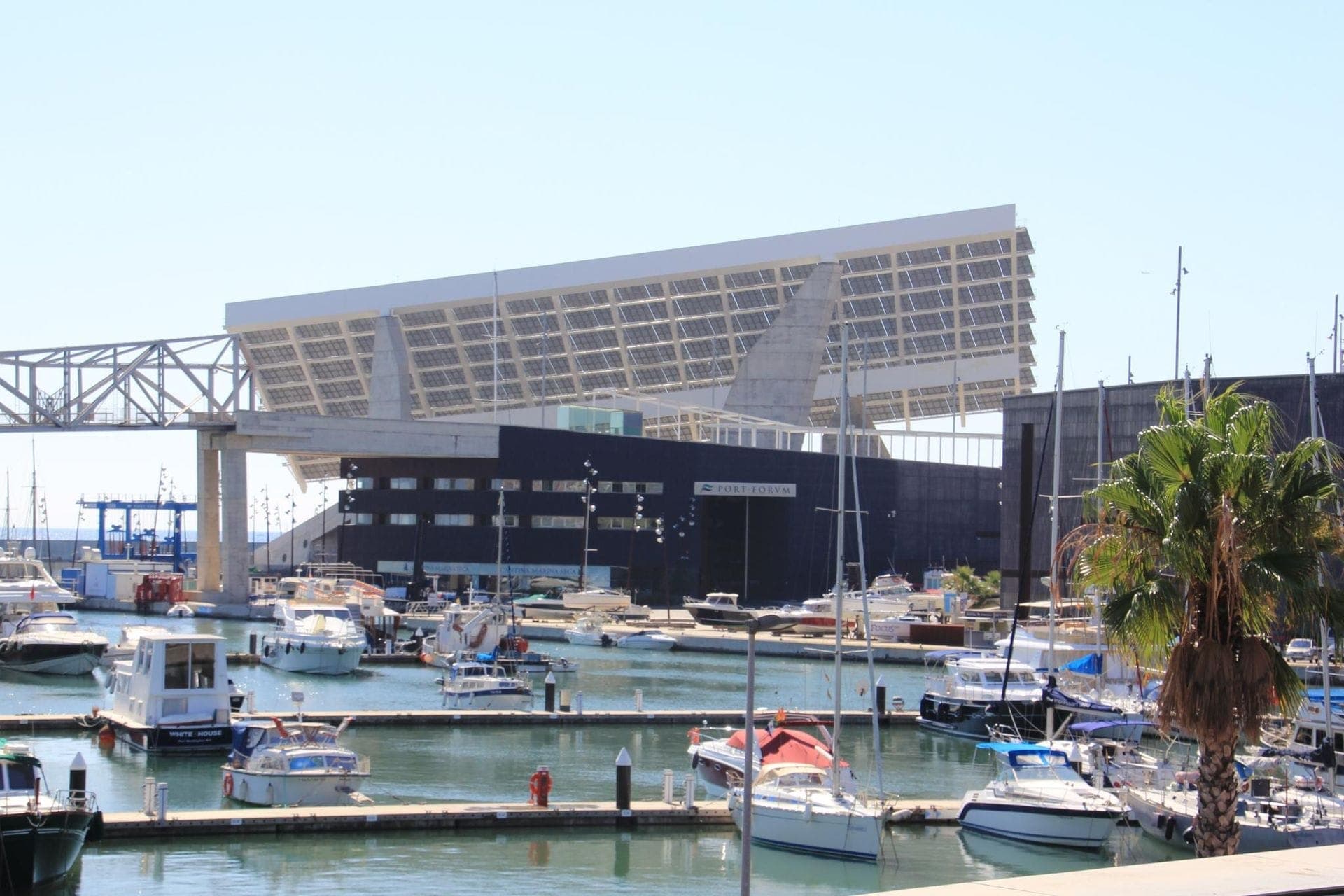 โครงสร้างแผงโซลาร์เซลล์สมัยใหม่ที่ Port Fòrum Barcelona พร้อมเรือยอชท์จอดเทียบท่าในมารีน่าที่สงบและท้องฟ้าสีฟ้า