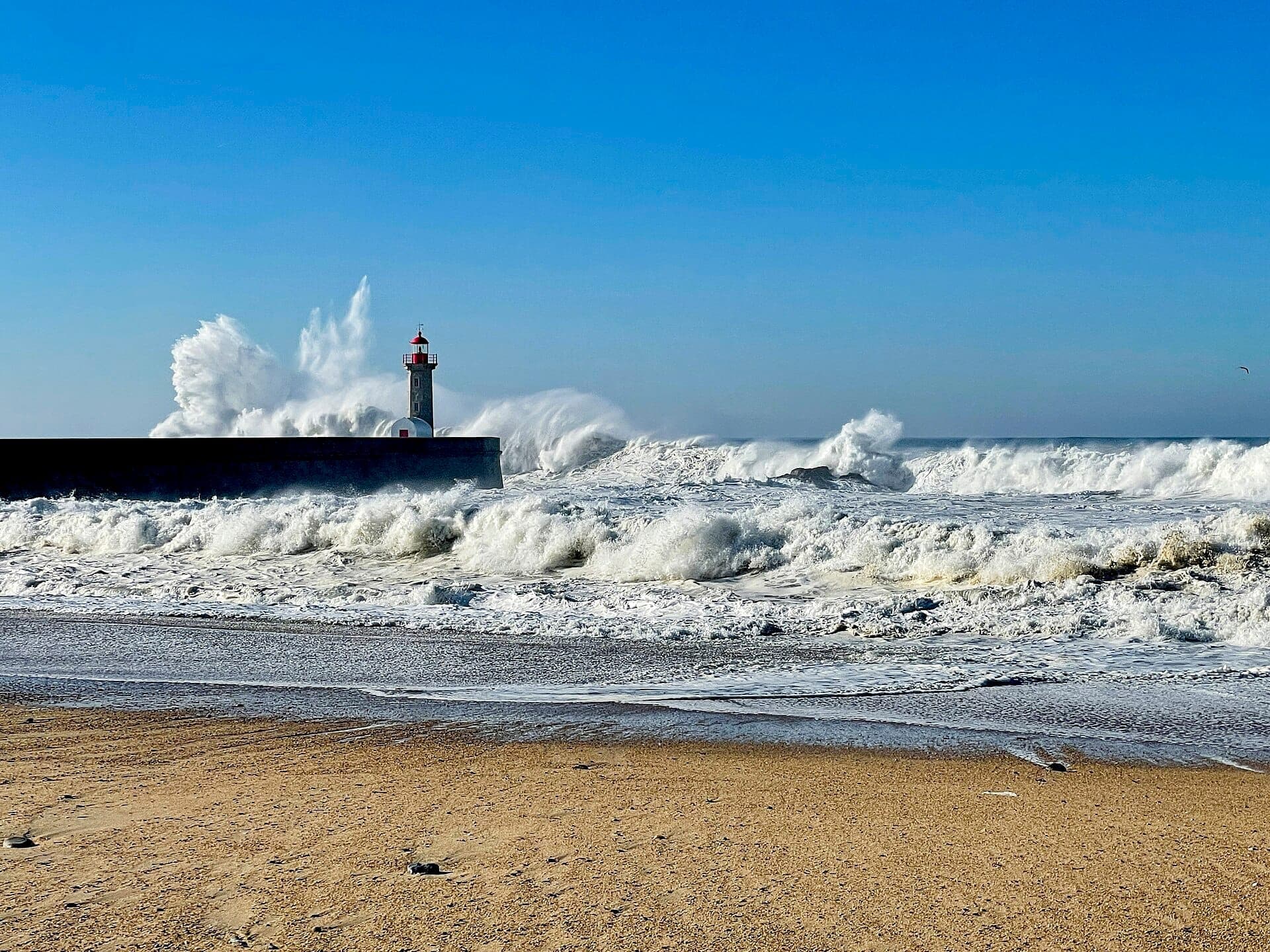 คลื่นซัดเข้าหาประภาคารอย่างน่าตื่นตาที่ชายหาด Foz do Douro พร้อมชายฝั่งทรายและท้องฟ้าสีฟ้าใสในเมือง Porto ประเทศโปรตุเกส