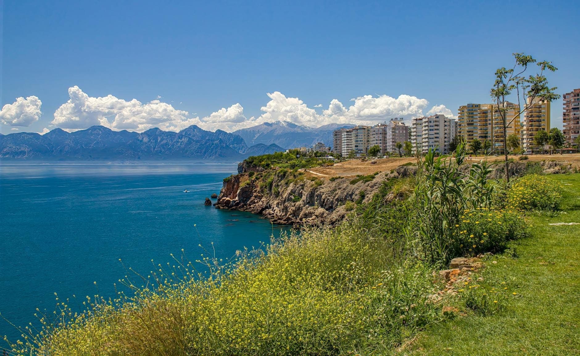 Klippenpark in Antalya mit grünem Gras, Wildblumen und Gebäuden mit Blick auf das Mittelmeer und ferne Berge.
