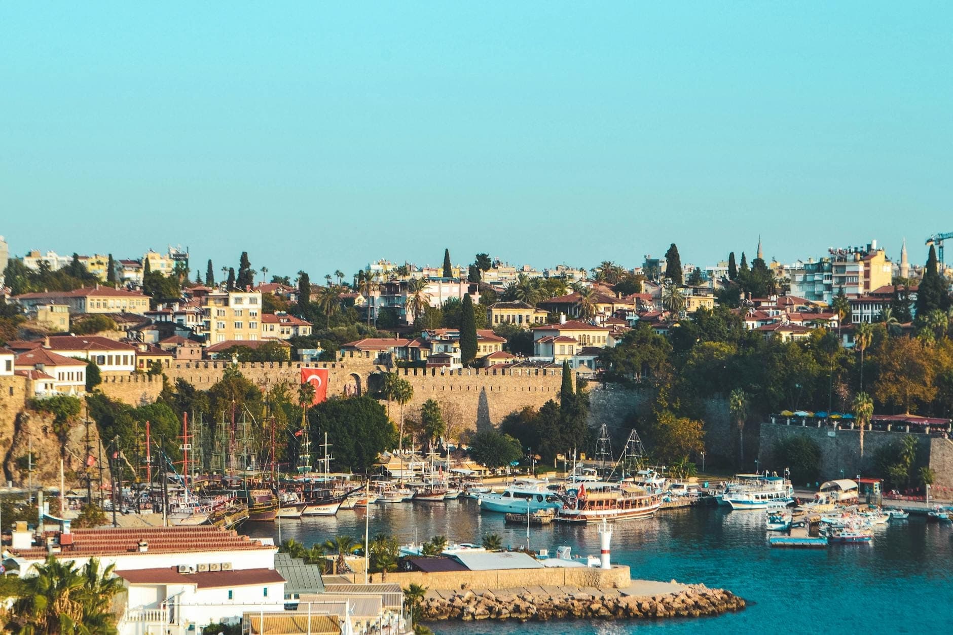 Panoramablick auf den Hafen der Altstadt von Antalya mit historischen Stadtmauern, bunten Booten auf blauem Wasser und mediterraner Architektur unter klarem Himmel.
