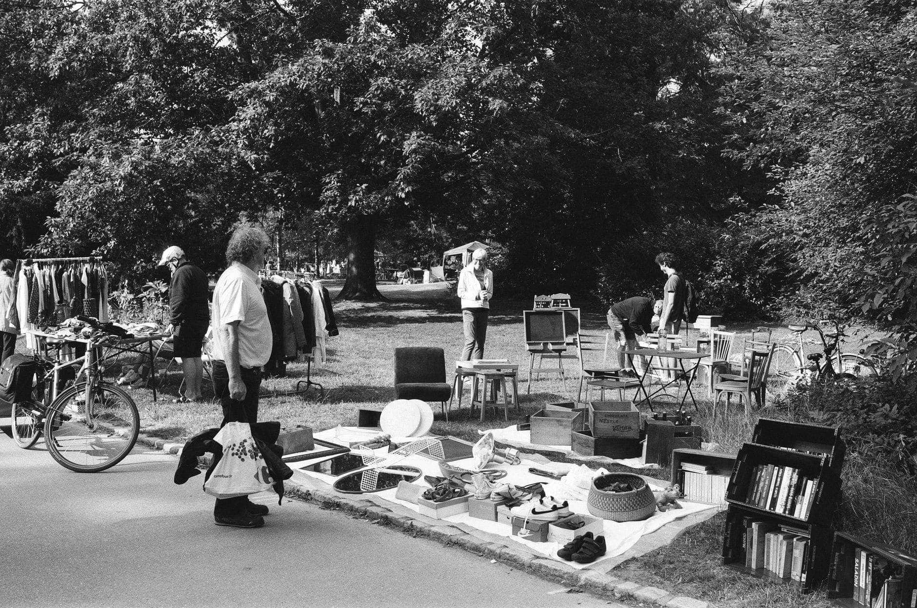Foto in bianco e nero di persone che curiosano tra gli articoli di un piccolo mercatino delle pulci all'aperto in un parco alberato, con alberi sullo sfondo.