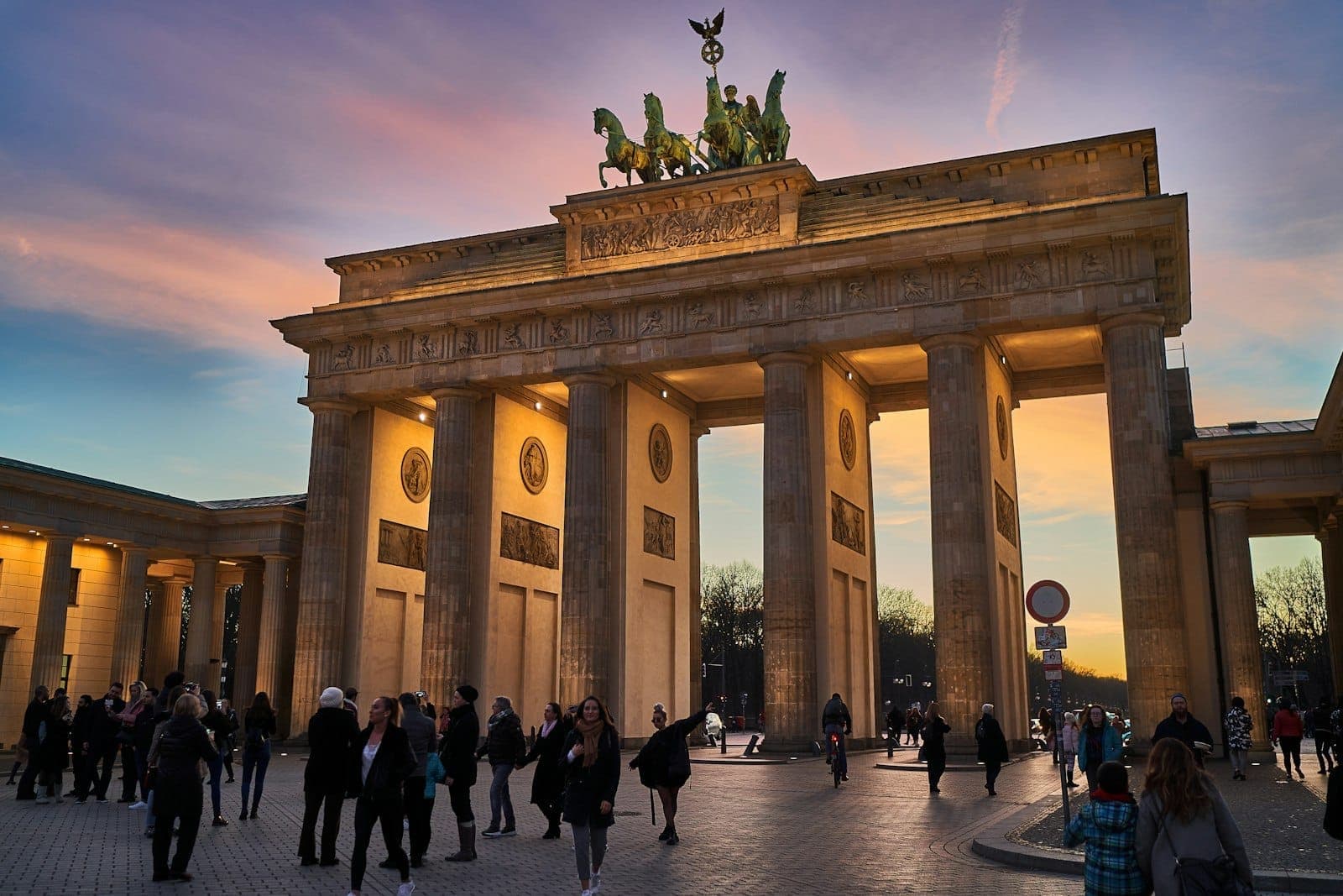 Folle di persone che camminano davanti alla illuminata Porta di Brandeburgo a Berlino al tramonto, con un cielo colorato sopra.