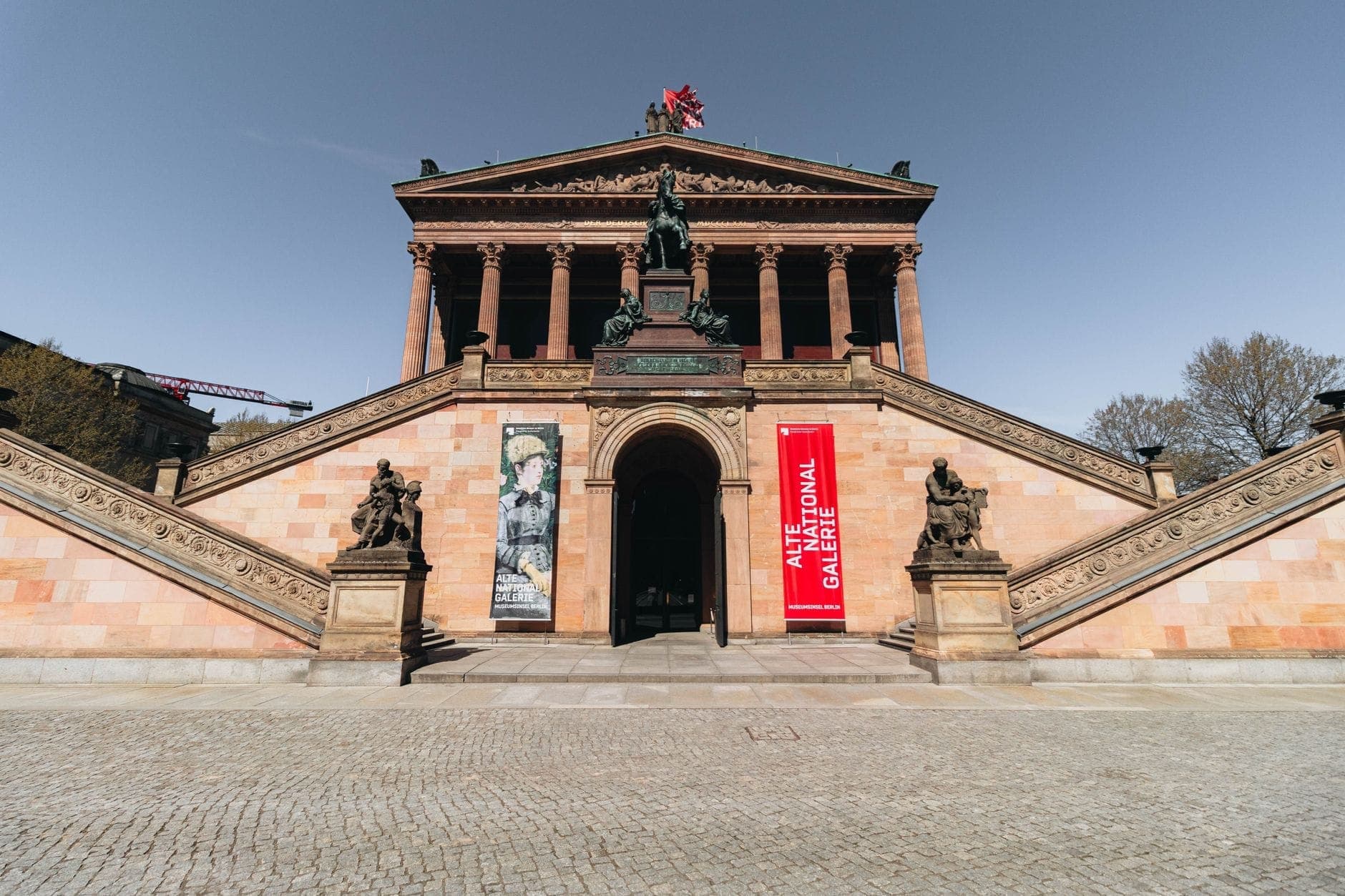 Grande ingresso di un museo di Berlino con ampie scale in pietra, statue, colonne e striscioni con la scritta Alte Nationalgalerie. Cielo limpido sopra l'edificio storico.