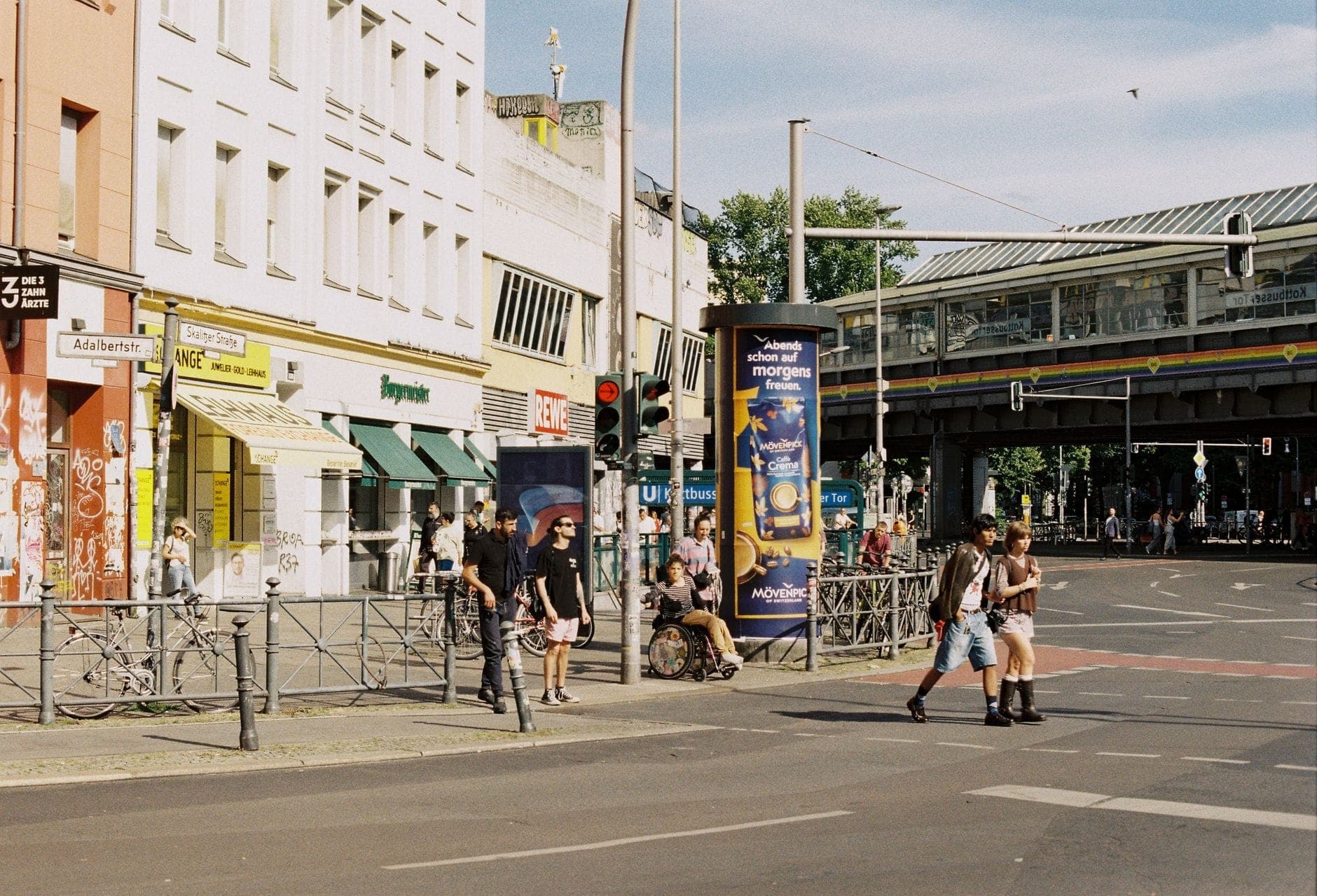 Un vivace angolo di strada a Berlino con persone che camminano e vanno in bicicletta, negozi, un ponte ferroviario e un'atmosfera di quartiere urbano durante il giorno.