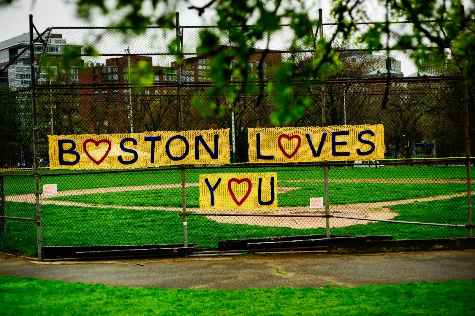 Terrain de baseball de Boston Common avec une clôture à mailles de chaîne décorée d'un panneau jaune fait main indiquant 'Boston Loves You' par une journée nuageuse.