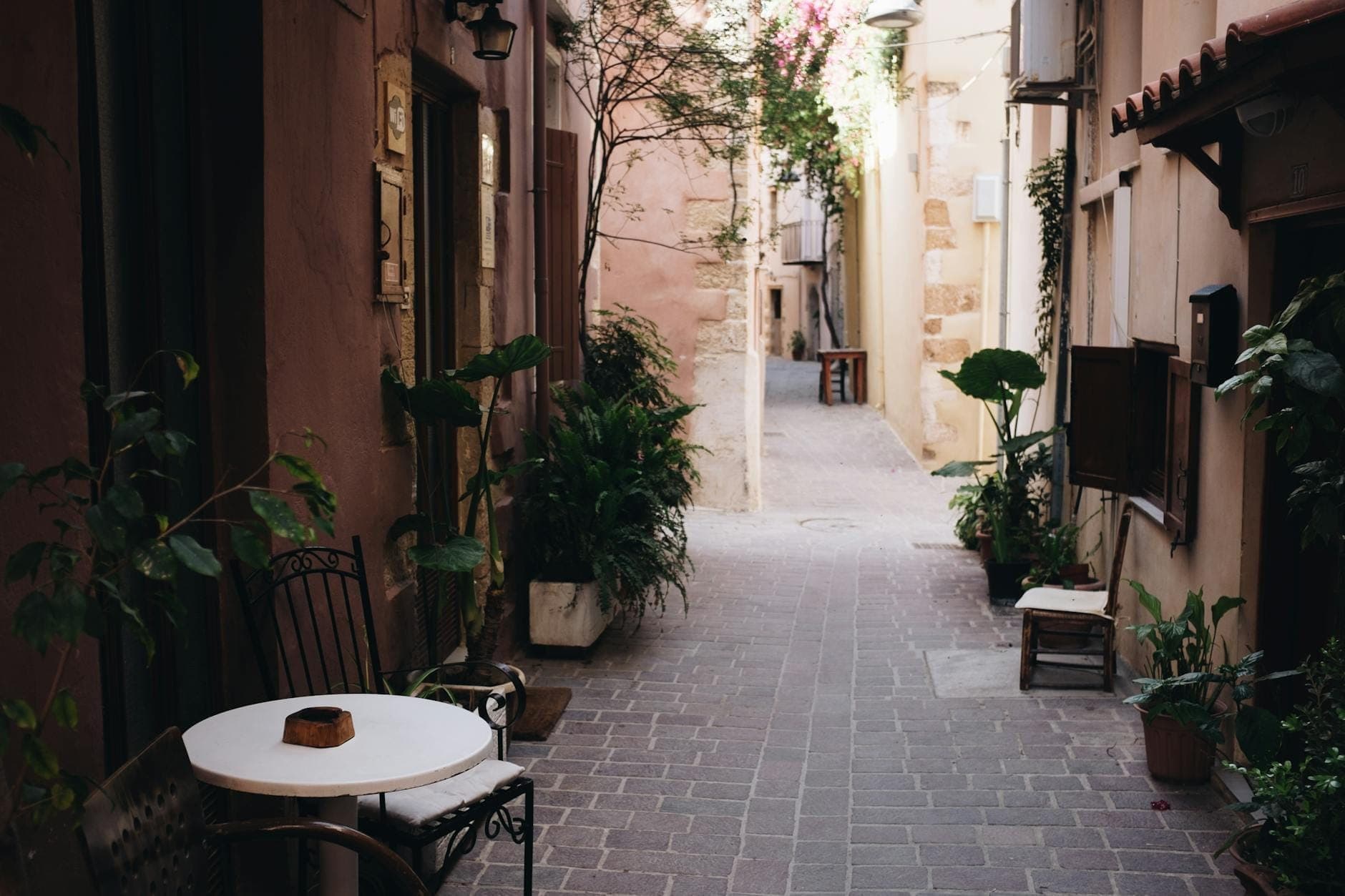 Stretto vicolo pedonale nella storica città vecchia di Creta con piante in vaso, un tavolo da caffè e edifici in pietra illuminati dal sole.