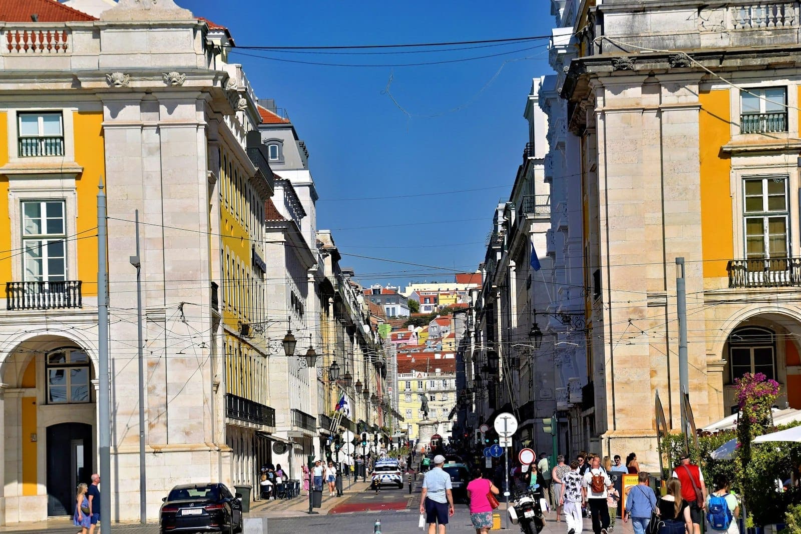 Vista ampla de uma movimentada rua de Lisboa com grandes edifícios históricos, pessoas caminhando e um vislumbre de casas coloridas ao fundo sob um céu azul limpo.