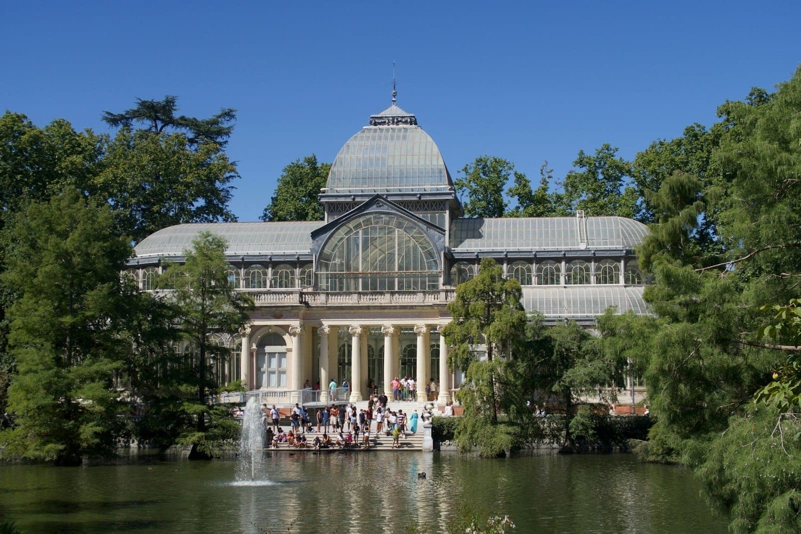 Vista del Palacio de Cristal di Madrid dall'altra parte di uno stagno, circondato da una rigogliosa vegetazione e visitatori che si godono il parco.