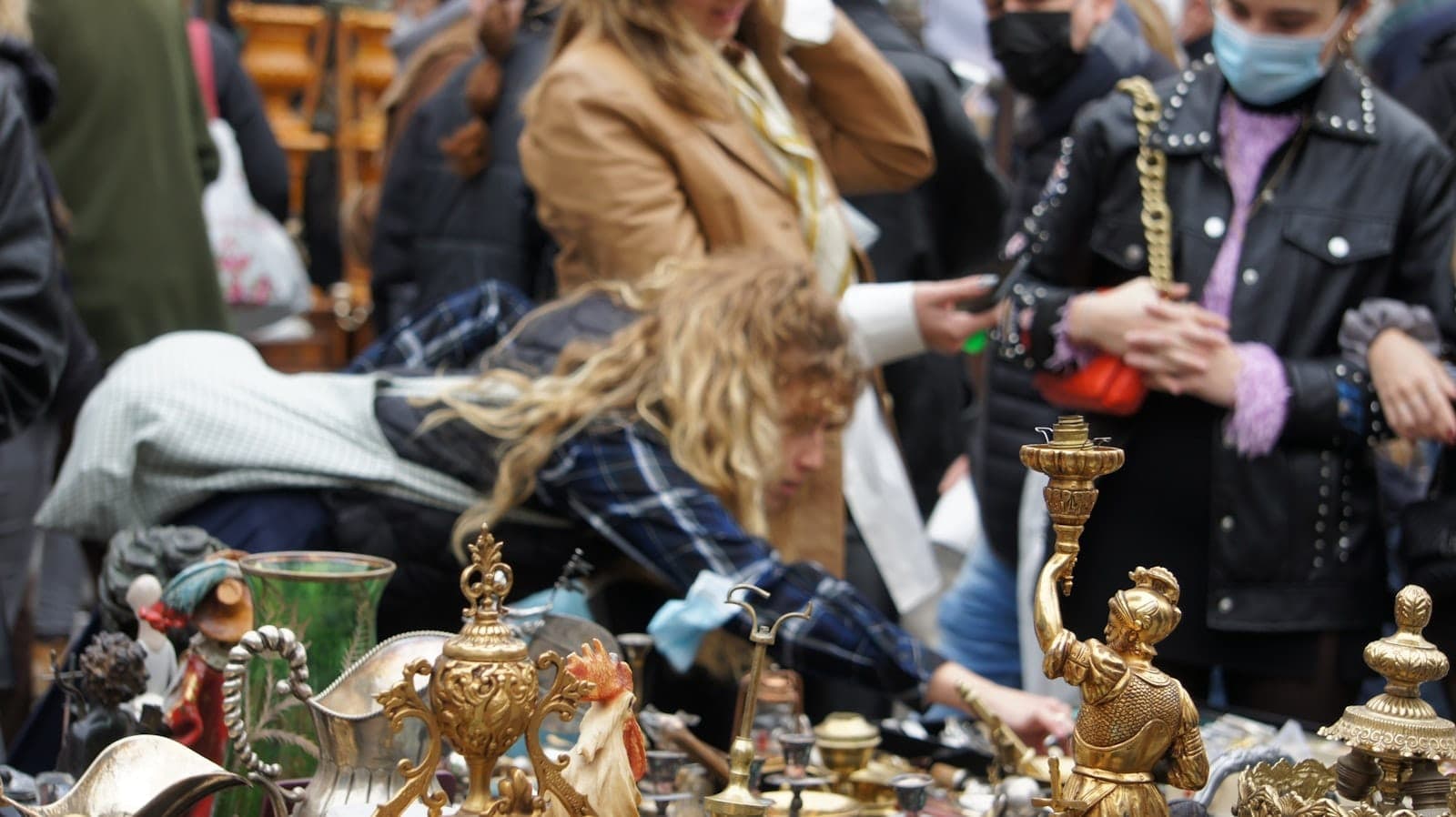 Persone che curiosano tra i banchi di un affollato mercato delle pulci all'aperto, esaminando oggetti vintage in ottone e antiquariato su un tavolo pieno.