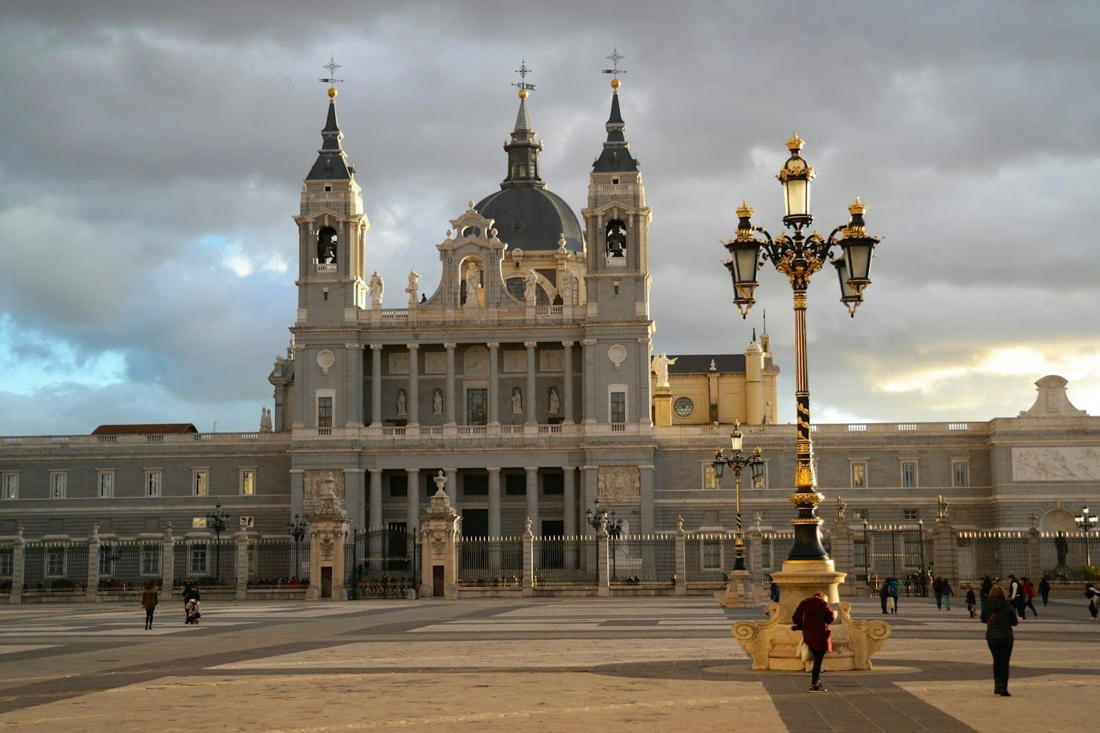 Vista ampia della Catedral de la Almudena di Madrid con le sue torri e la cupola centrale, vista da una piazza con lampioni ornati e persone che camminano.