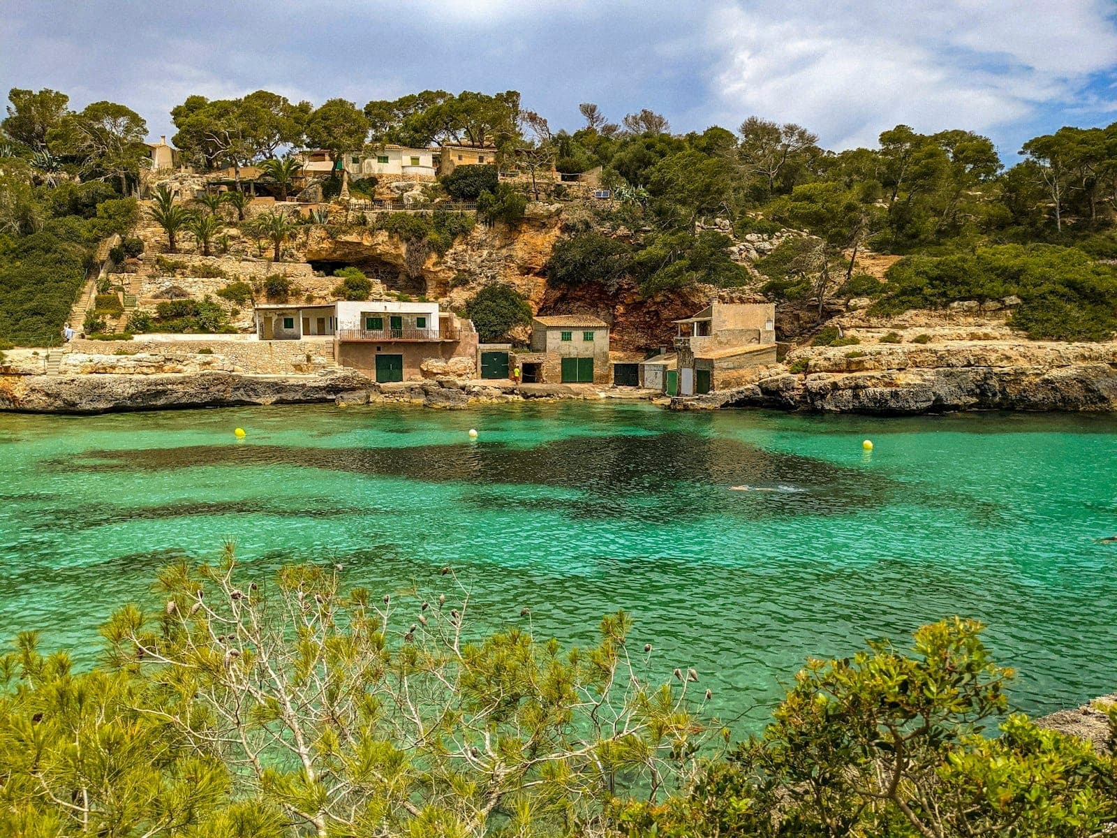 Malerische Bucht auf Mallorca mit türkisblauem Wasser, traditionellen Fischerhäusern auf Felsvorsprüngen und üppigen Bäumen unter strahlend blauem Himmel.