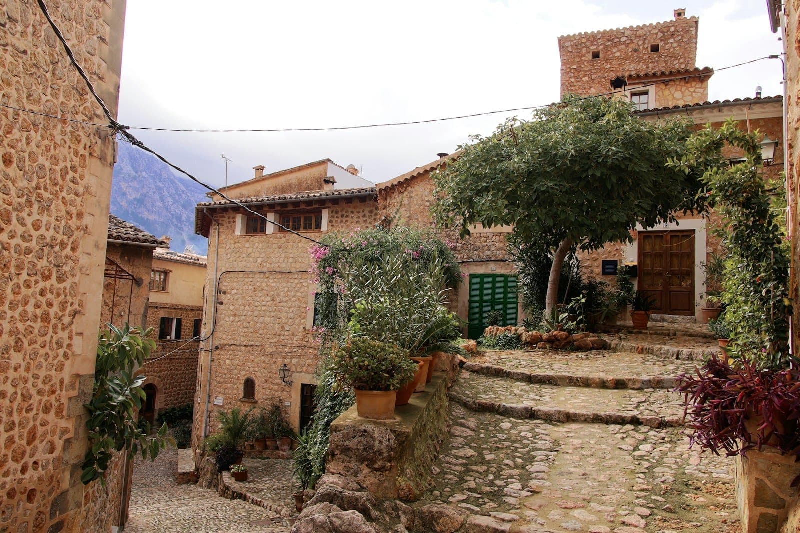 Steinstufen und gepflasterter Weg, der sich zwischen alten Steinhäusern und Grünflächen in einem historischen Dorf auf Mallorca schlängelt.