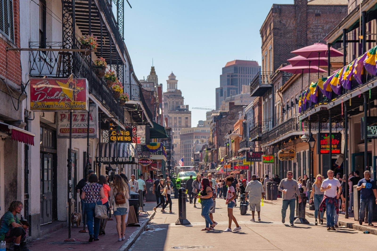 People walking along a lively street in the French Quarter of New Orleans, with historic buildings, balconies, and colorful Mardi Gras decorations.