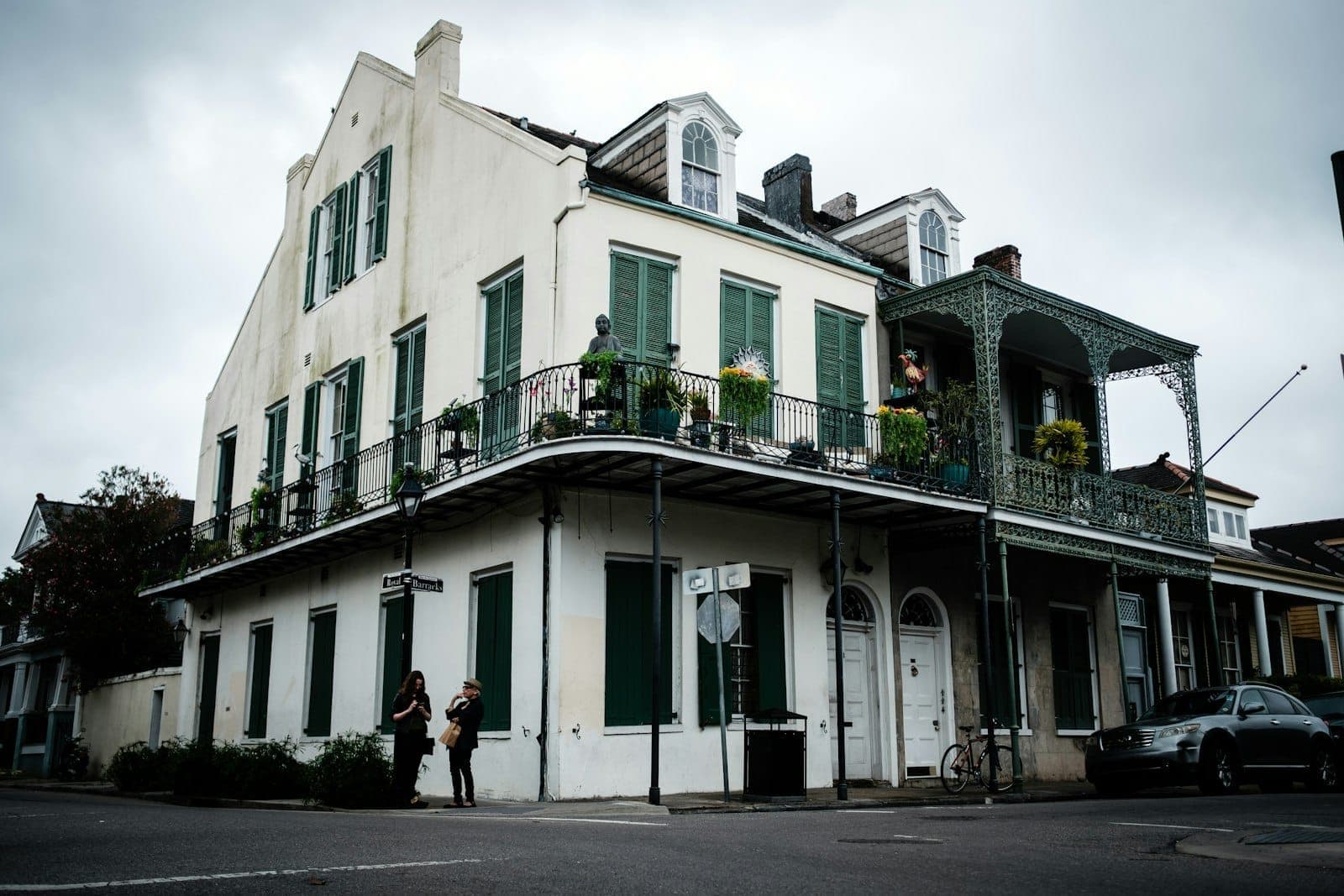 Narożnik ulicy w New Orleans z klasycznym budynkiem w dzielnicy French Quarter, zdobionym kutym balkonem, zielonymi okiennicami i przechodniami poniżej.