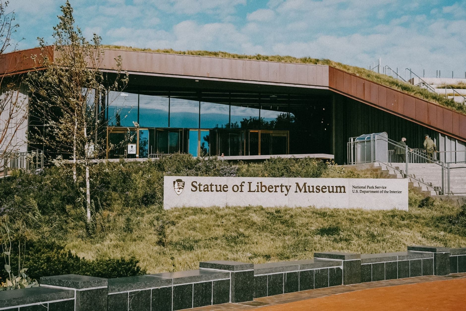ทางเข้าภายนอกของ Statue of Liberty Museum พร้อมป้ายและผนังกระจกสมัยใหม่ ล้อมรอบด้วยพืชพรรณเขียวขจี