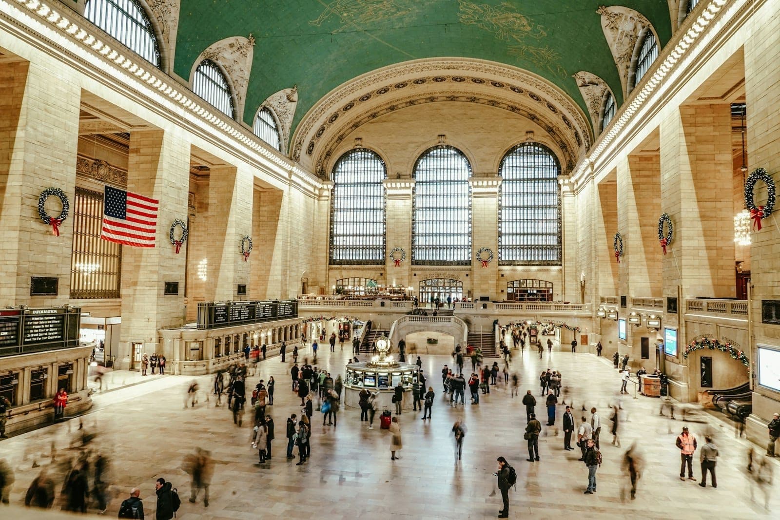 ล็อบบี้หลักของ Grand Central Terminal ที่เต็มไปด้วยผู้คน พร้อมหน้าต่างโค้งอันเป็นเอกลักษณ์ เพดานลวดลายท้องฟ้า และธงชาติอเมริกันที่มองเห็นได้ชัดเจน