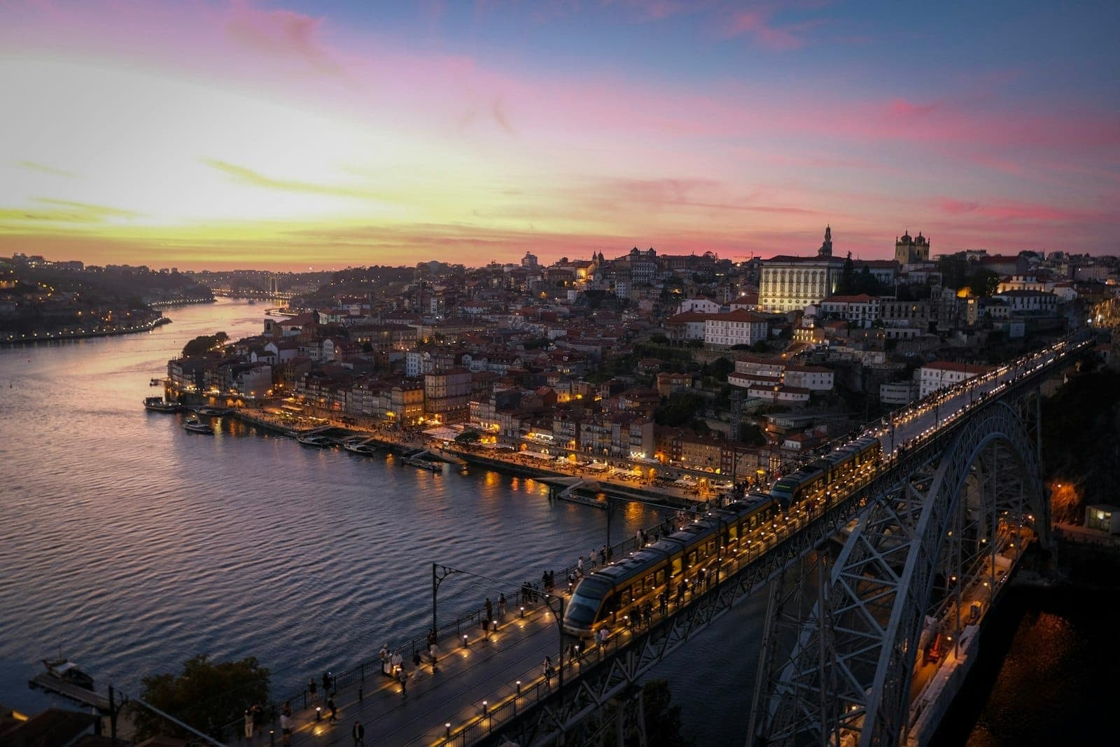 เส้นขอบฟ้าของเมืองปอร์โตยามพระอาทิตย์ตกพร้อมสะพาน Dom Luís I และแม่น้ำ Douro ถนนที่สว่างไสวและทางเดินริมน้ำตามแนวอาคารเมืองที่มีชีวิตชีวา