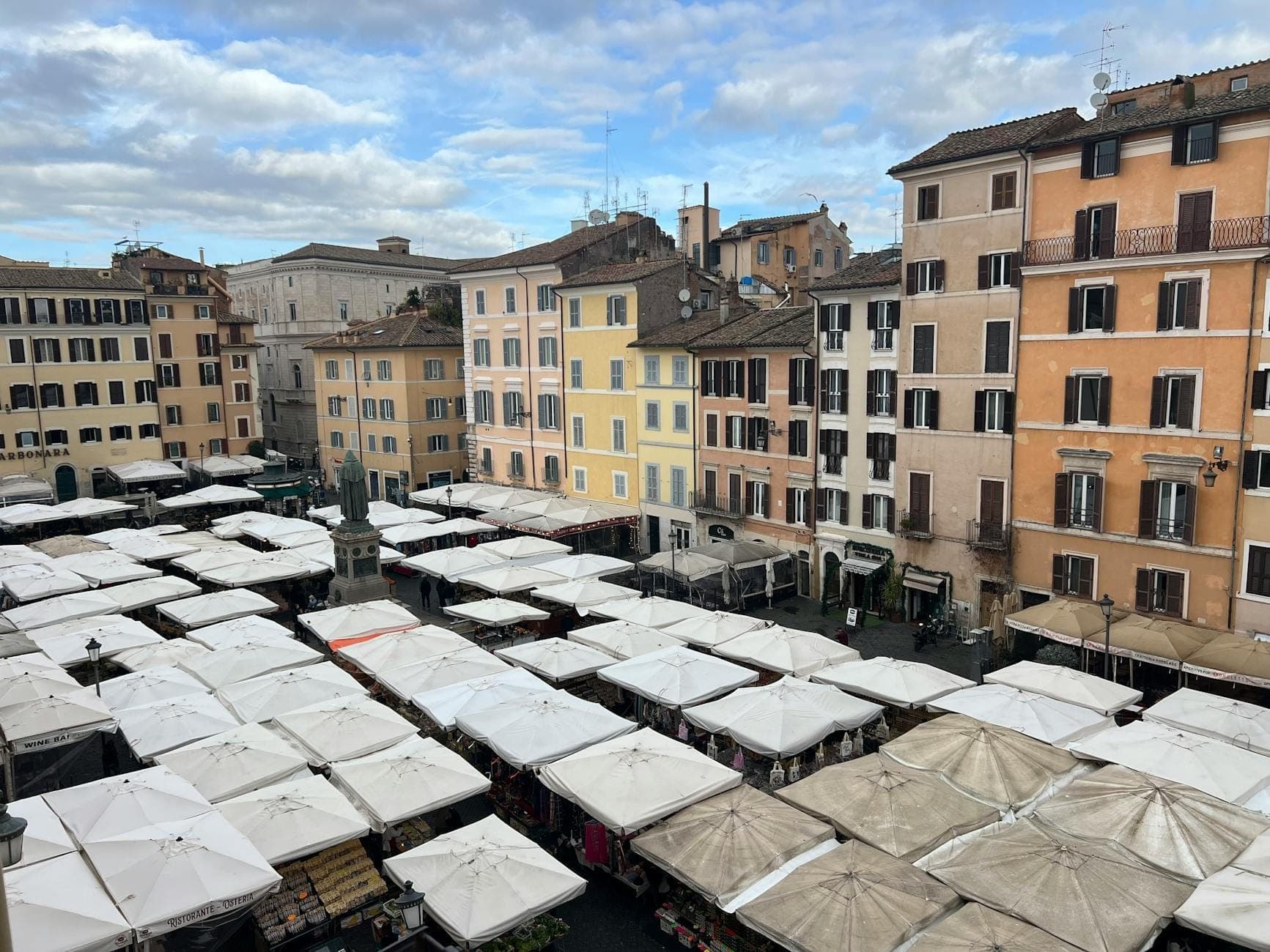 Une place romaine animée avec des dizaines d'étals de marché sous des auvents blancs, entourée d'immeubles d'appartements anciens colorés et d'un ciel ouvert.