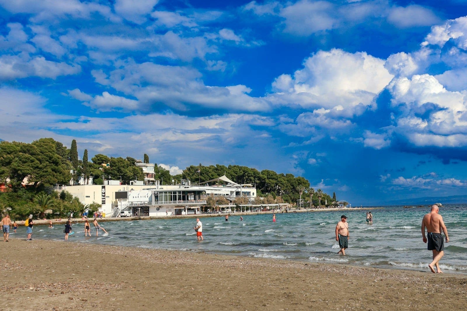 Des personnes se baignant et se relaxant sur une plage de sable à Split, en Croatie, avec des bâtiments, des arbres et un ciel bleu vibrant en arrière-plan.