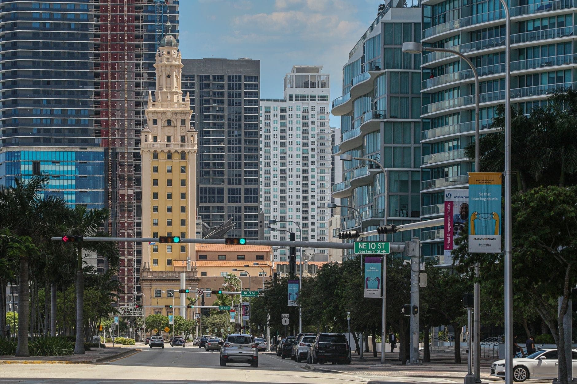 Freedom Tower prominently centered among modern Miami skyscrapers, viewed down a bustling city street under a bright sky, with traffic and palm trees framing the scene.