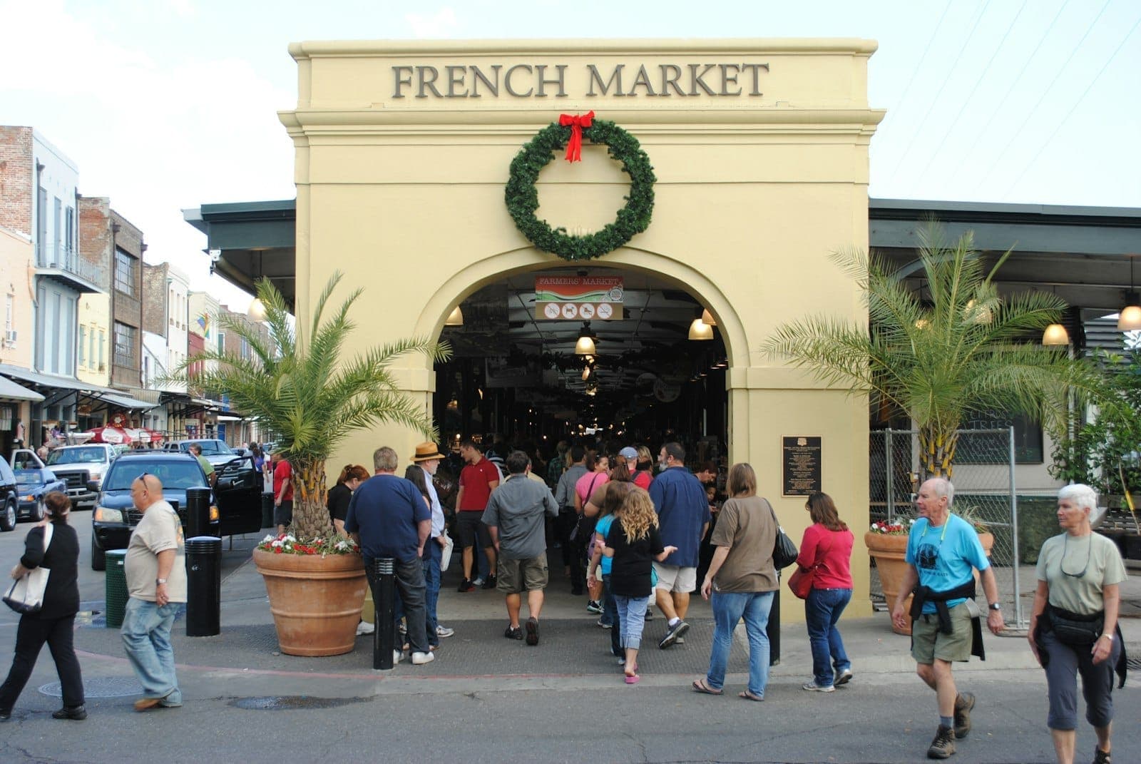 Entrance to the French Market in New Orleans with a holiday wreath, palm trees, and crowds of people walking by and inside.