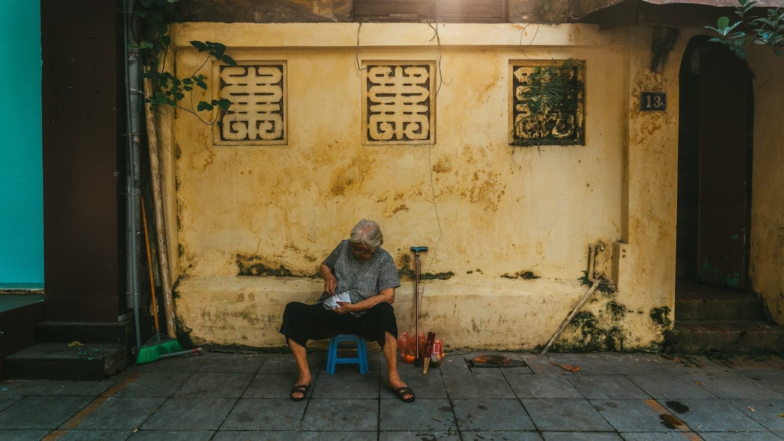 Starsza osoba siedzi na niebieskim stołku przy ochrowej ścianie z francuskimi detalami architektonicznymi, oddając spokojną atmosferę Dzielnicy Francuskiej w Hanoi.