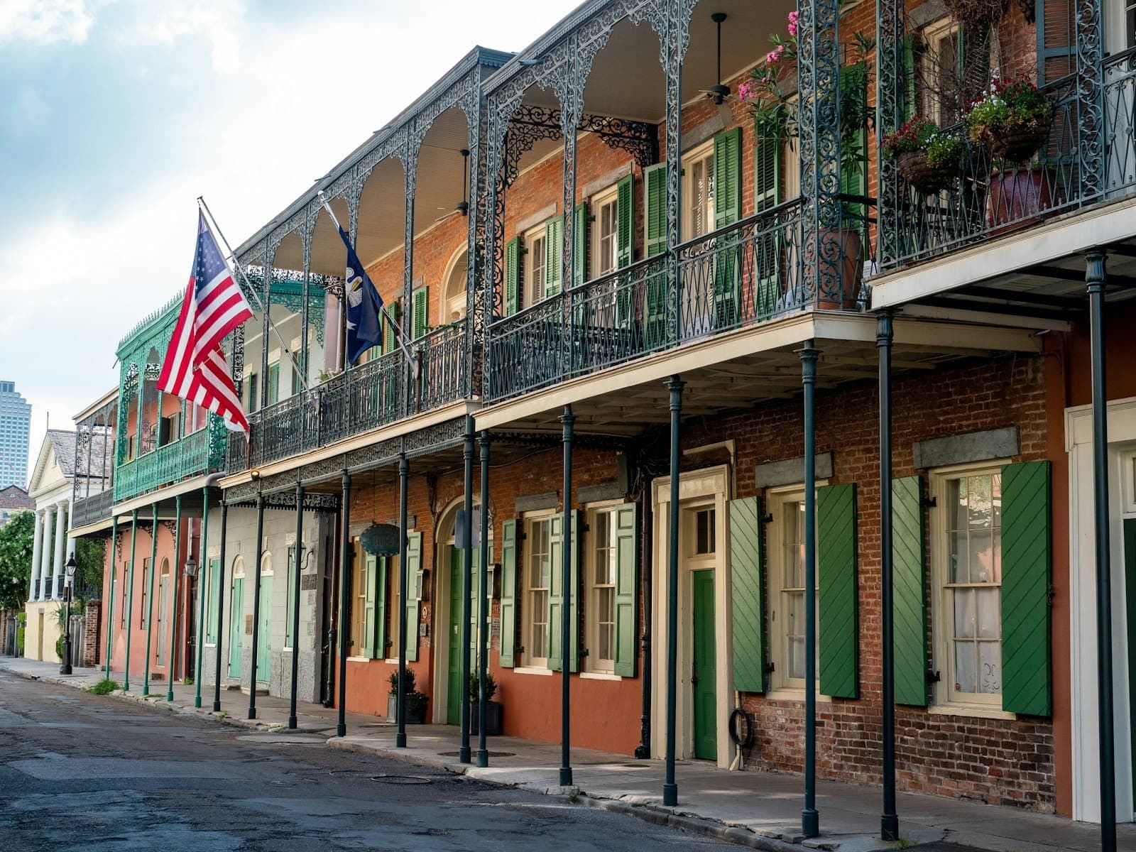 Classic French Quarter street view with colorful buildings, ornate iron balconies, green shutters, American flags, and a quiet sidewalk under soft daylight.