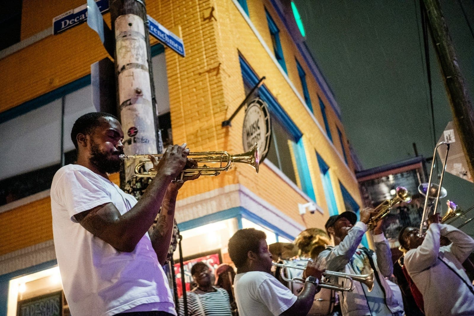 Músicos tocam instrumentos de sopro na Frenchmen Street à noite, com um edifício de tijolos amarelos e placas de rua visíveis sob as luzes da cidade.
