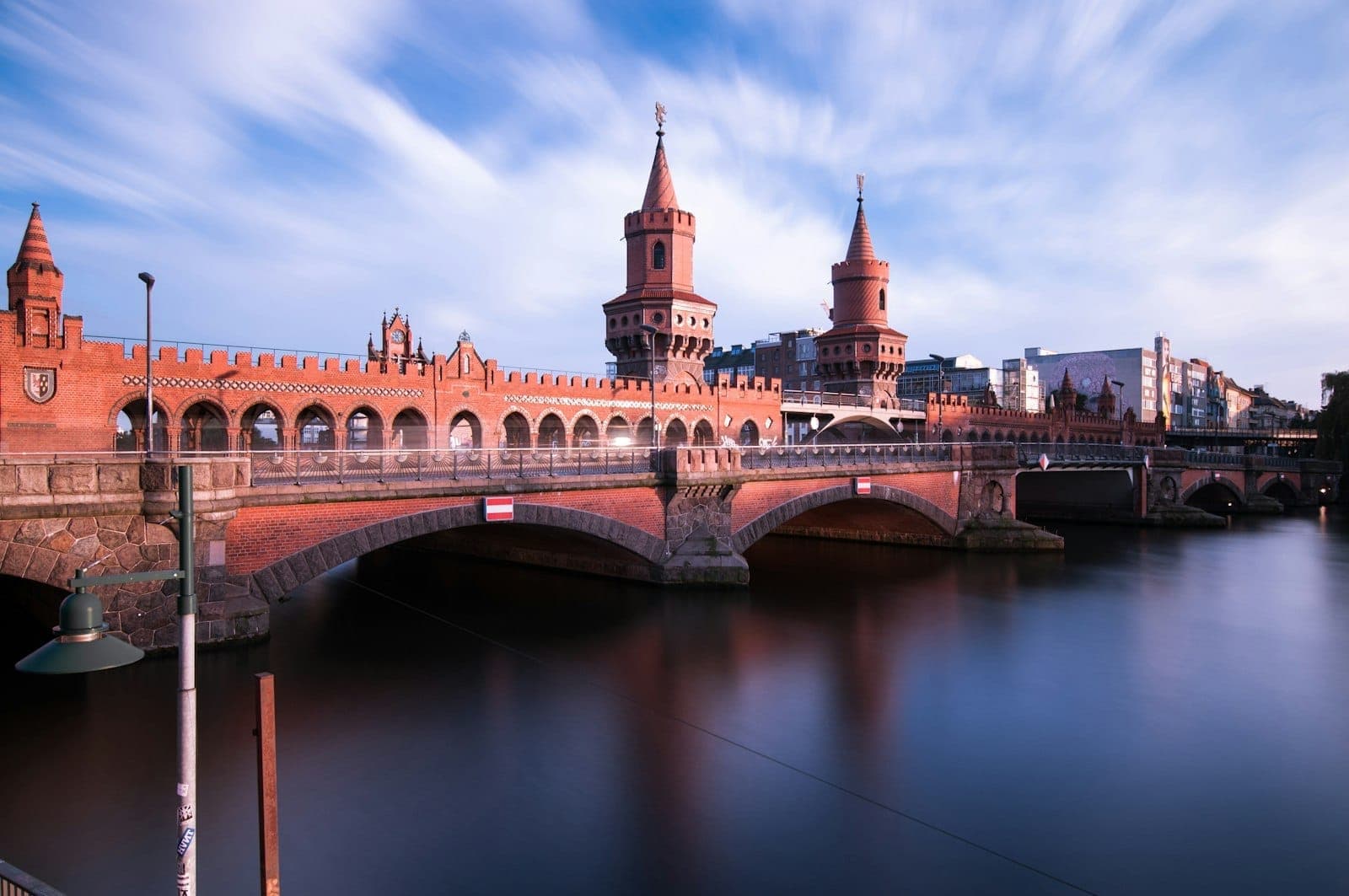 สะพาน Oberbaum ในย่าน Friedrichshain, เบอร์ลิน แดดหวานยามเย็น ฉากหลังเป็นหอคอยอิฐสีแดงสะท้อนในแม่น้ำ Spree กับก้อนเมฆแปรปรวนเหนือศีรษะ