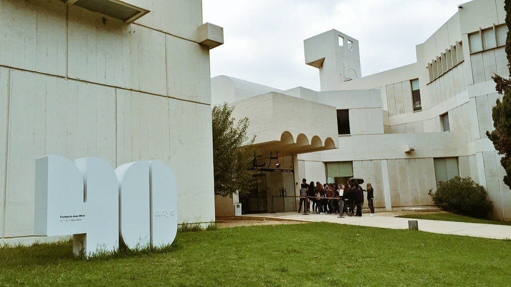มุมมองภายนอกของ Fundació Joan Miró ในบาร์เซโลนา แสดงให้เห็นสถาปัตยกรรมคอนกรีตสีขาวสมัยใหม่ สนามหญ้าสีเขียว และกลุ่มนักท่องเที่ยวบริเวณทางเข้า