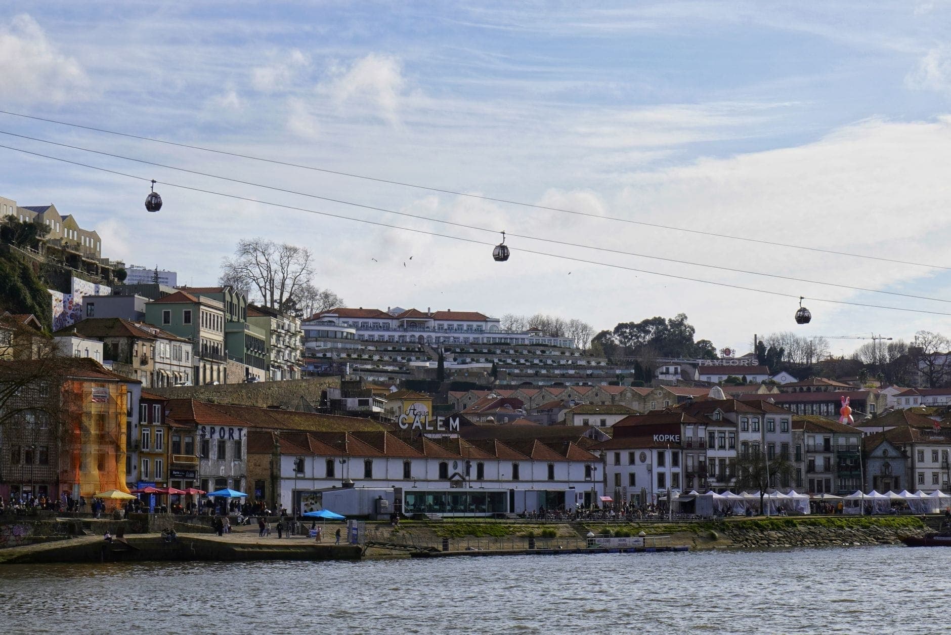 Gondeln schweben über das Flussufer von Vila Nova de Gaia in Richtung Dom-Luís-I-Brücke, mit historischen Gebäuden entlang des Douro unter blauem Himmel.