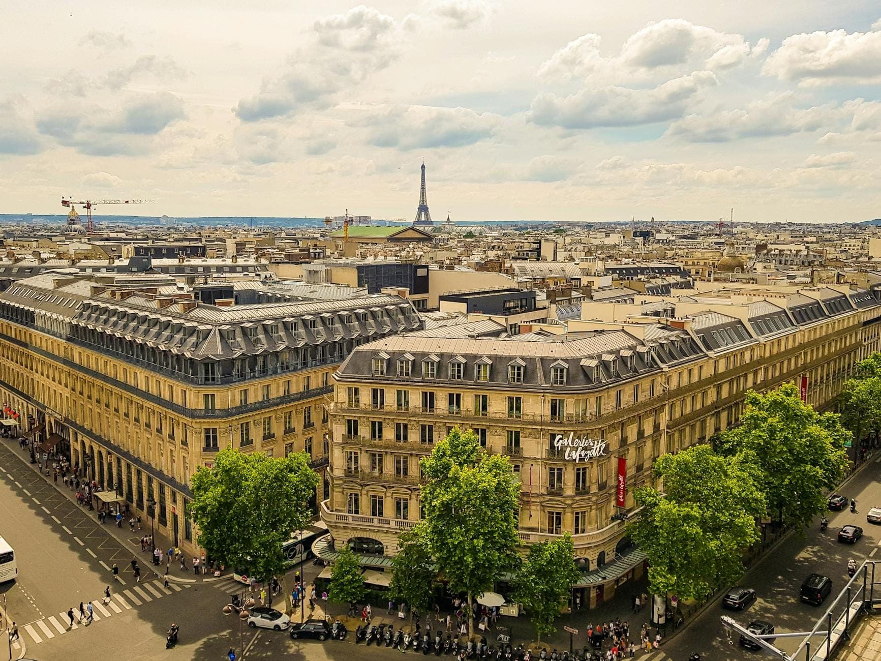 Wide aerial view of Galeries Lafayette Haussmann exterior with classic Parisian architecture, tree-lined street, and the Eiffel Tower in distance under a dramatic sky.