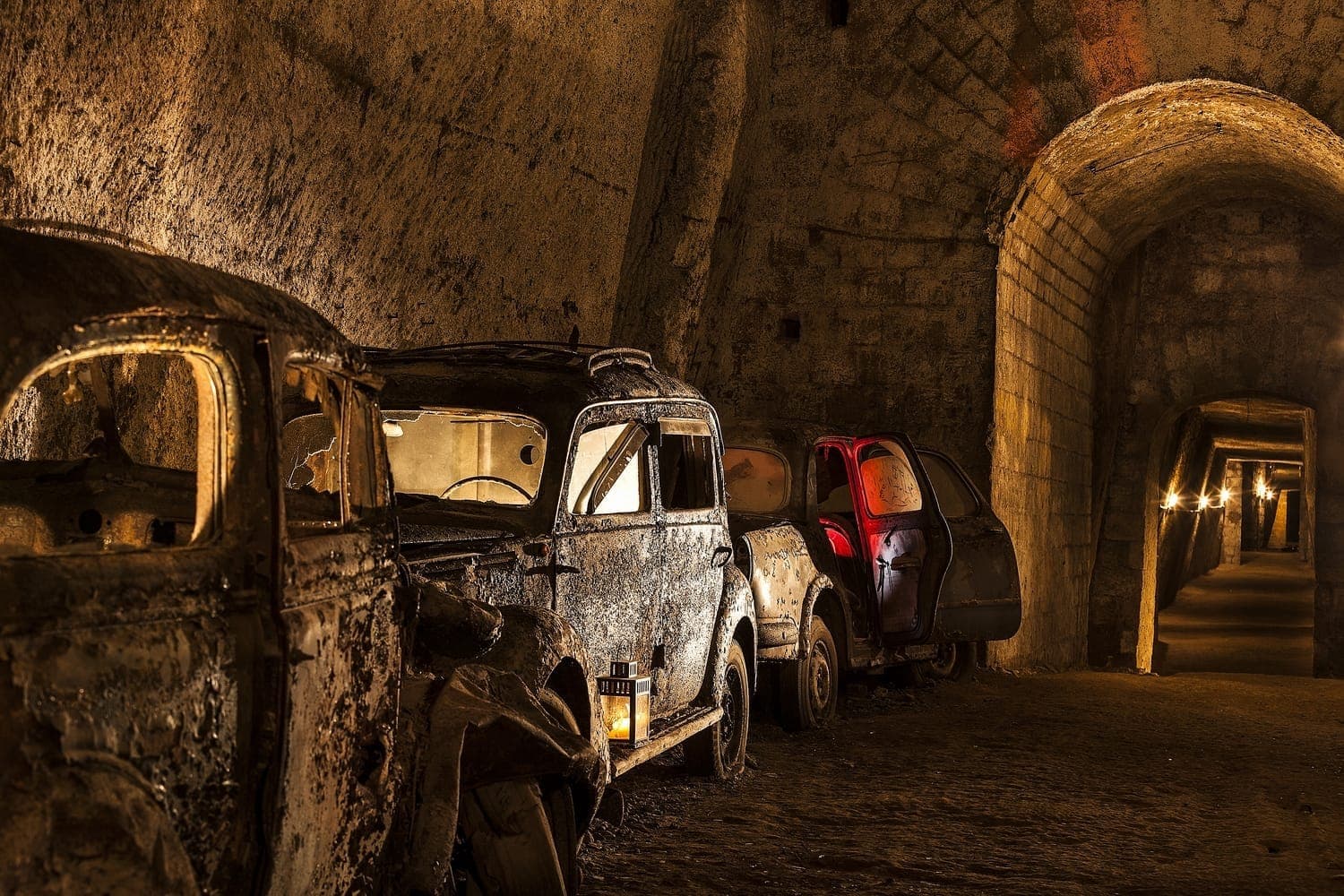 Carros enferrujados e abandonados enfileirados no túnel de pedra mal iluminado dentro da Galleria Borbonica, com arcos e paredes de rocha de tufo texturizadas à vista.