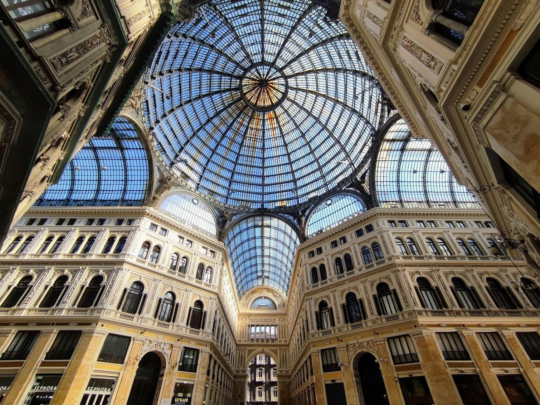 Vista em grande angular da majestosa cúpula de vidro da Galleria Umberto I e sua ornamentada arquitetura do século XIX, com a luz do sol atravessando os impressionantes tetos abobadados acima das elegantes galerias.