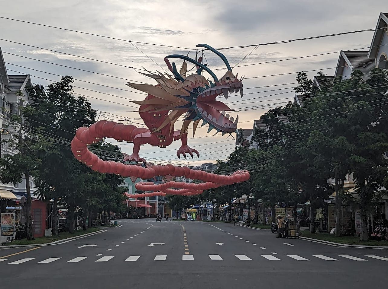 Malalaking pulang dragon na parol na nakabitin sa itaas ng kalye ng Grand World Phu Quoc na may palmerang hanay tuwing kapistahan, may makukulay na tindahan at tradisyonal na arkitekturang Vietnamese sa ilalim ng maulap na langit.