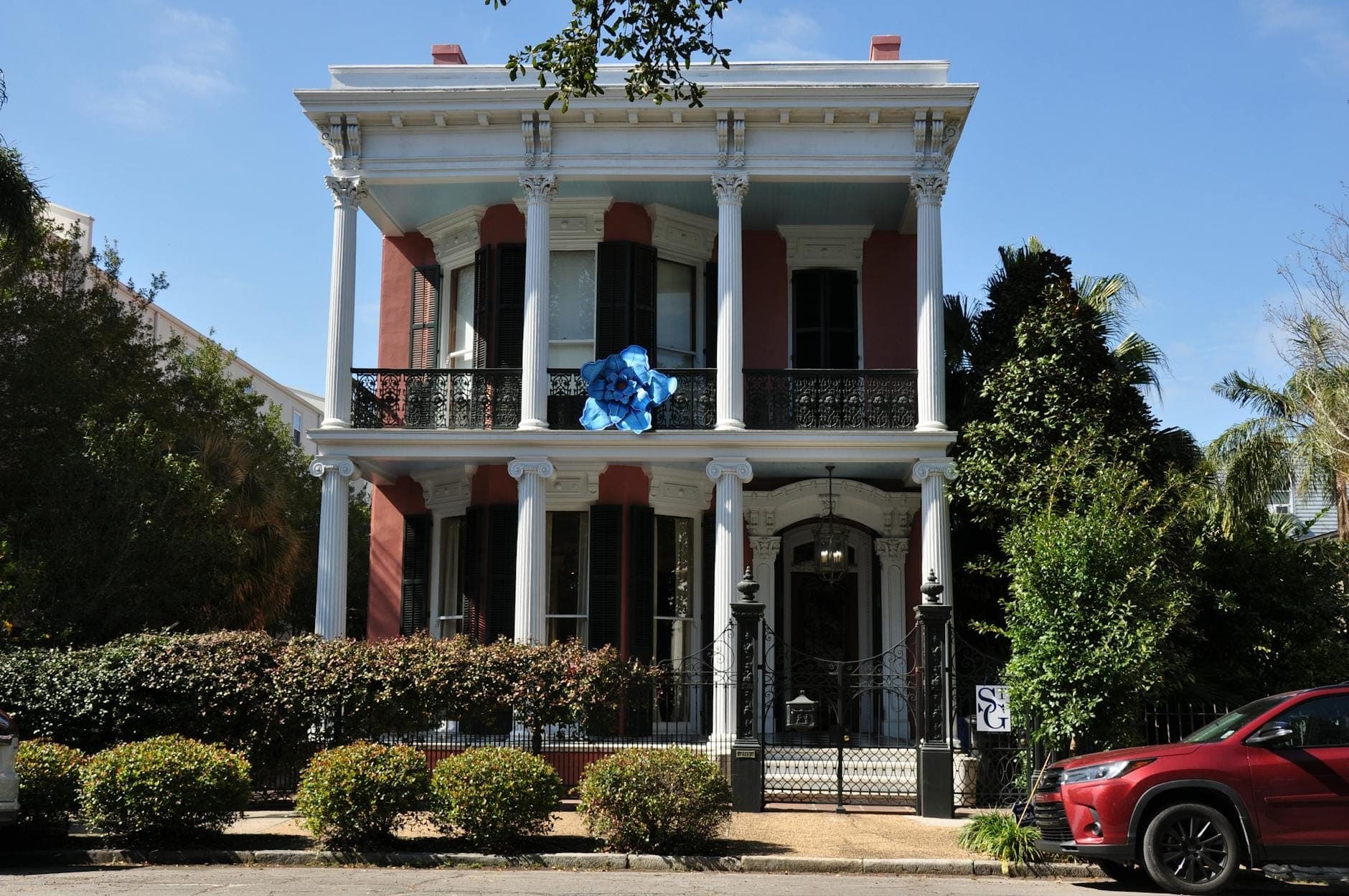A grand, historic mansion in the Garden District of New Orleans, featuring tall white columns, wrought-iron balcony railings, and lush greenery in front.
