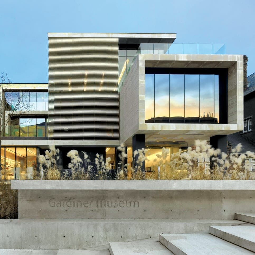 Esterno moderno del Gardiner Museum con architettura in vetro e pietra, erbe ornamentali in primo piano e una morbida luce serale che illumina l'edificio.