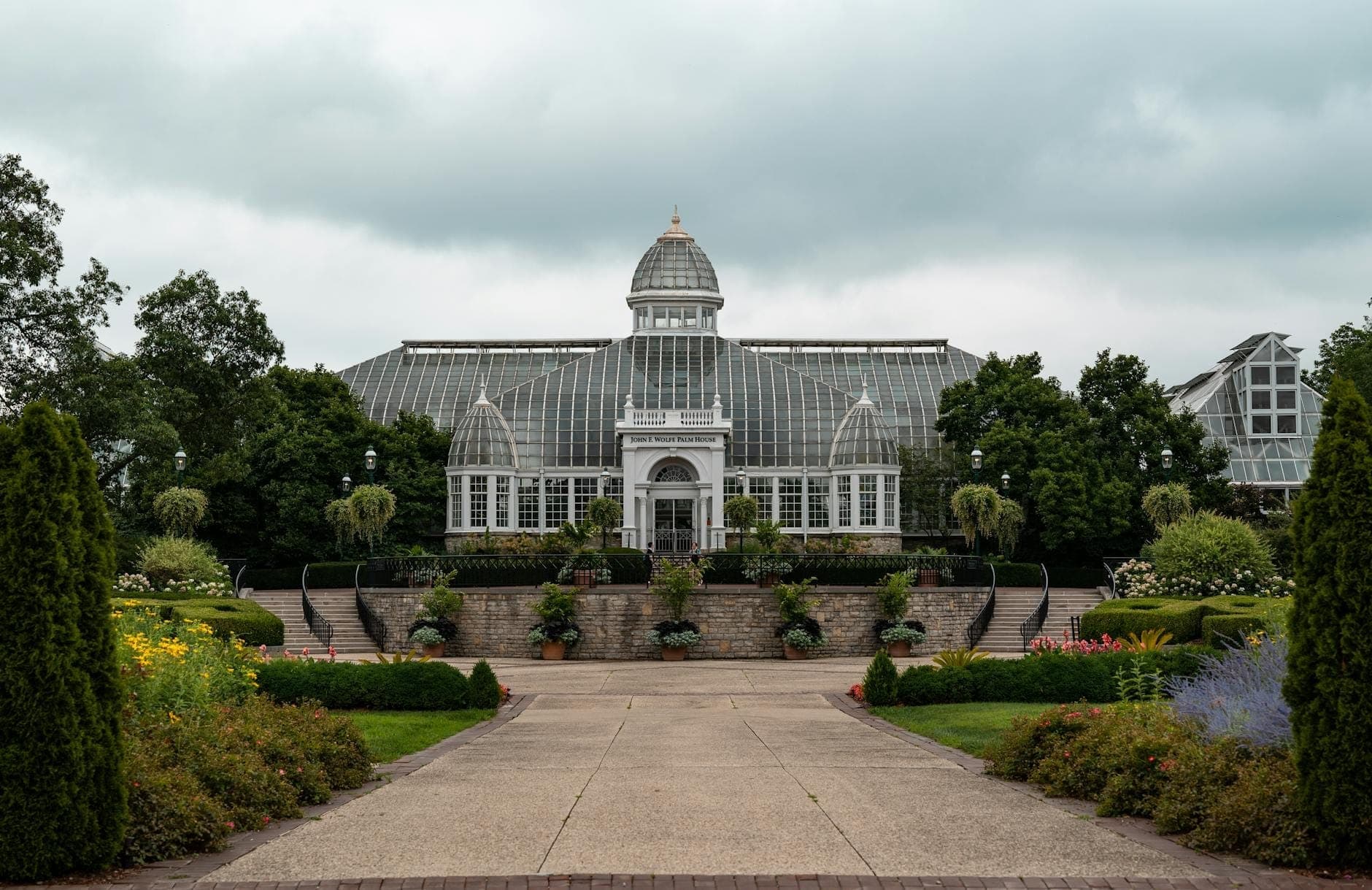 Vista amplia de la entrada del Garfield Park Conservatory y su cúpula de cristal, enmarcada por jardines exuberantes y un camino bajo un cielo dramático y nublado.