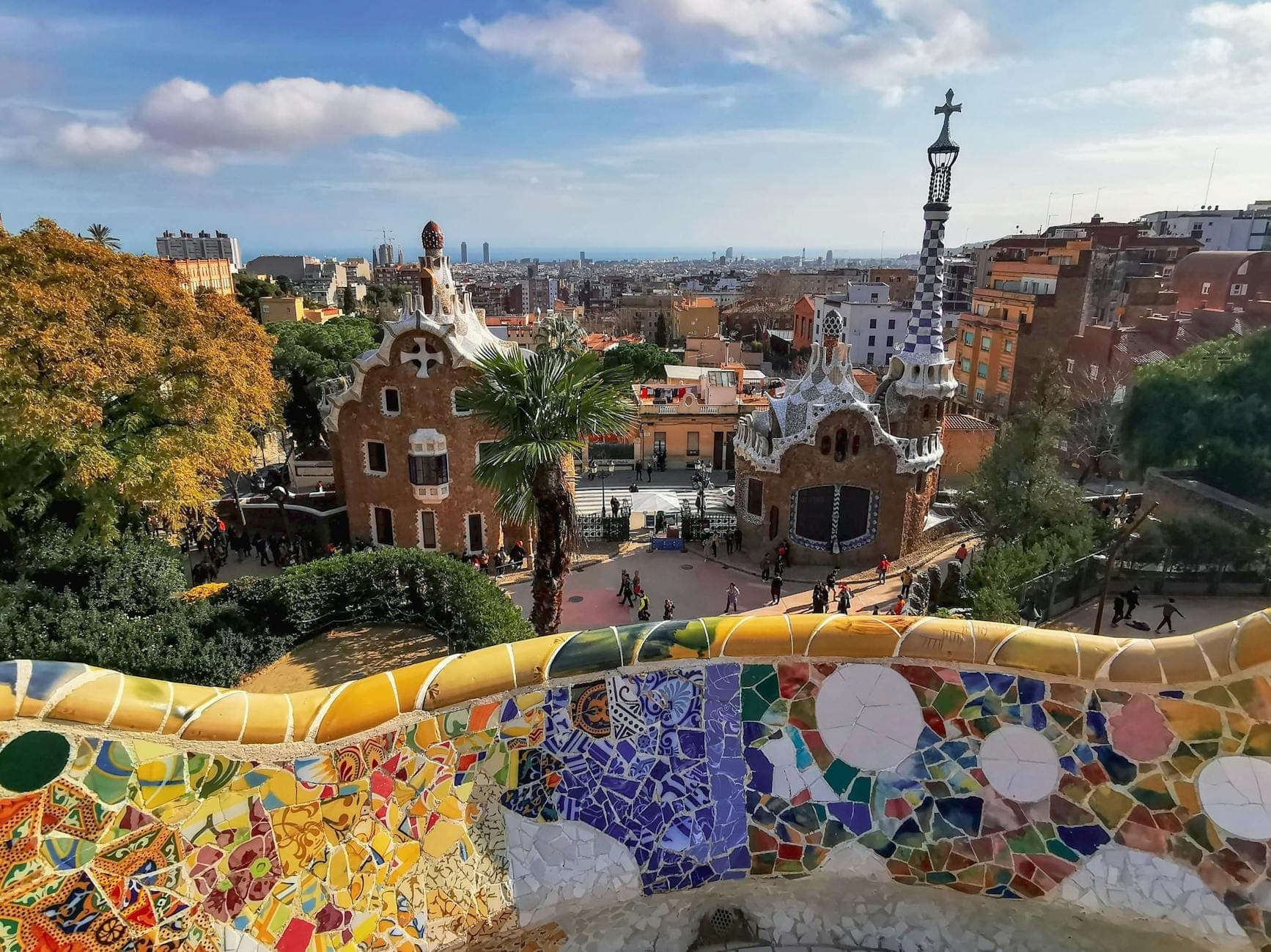 Pemandangan teras mosaik berwarna-warni Park Güell, rumah gerbang rancangan Gaudí, dan lanskap kota Barcelona di bawah langit biru.