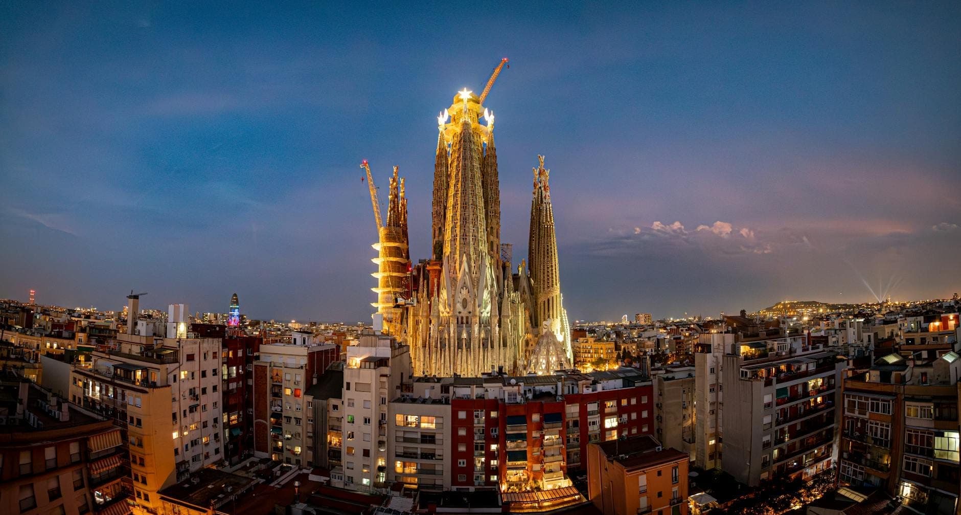 Pemandangan malam panoramik Sagrada Família yang diterangi cahaya menjulang di atas lanskap kota Barcelona, dengan derek konstruksi yang terlihat dan langit berwarna ungu di atasnya.