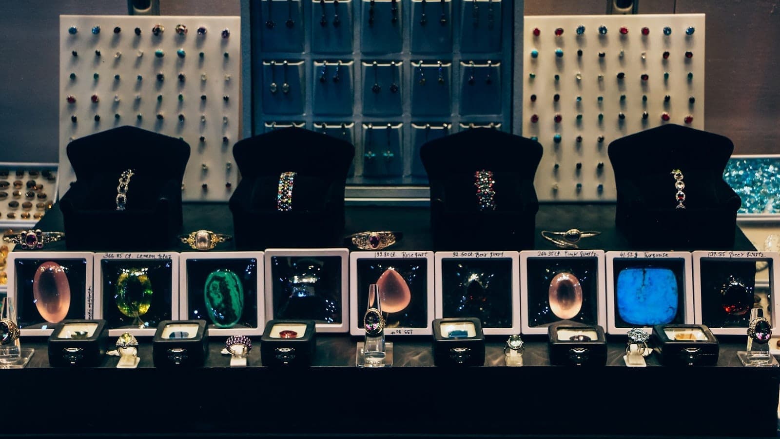 Display case at Gems Gallery Pattaya featuring colorful gemstones, rings, and bracelets under bright lighting with a backdrop of pinned jewelry samples.