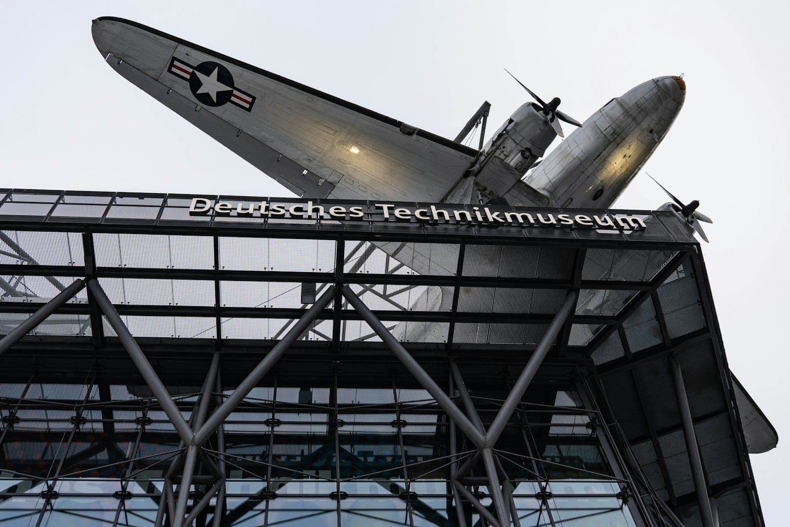 ภายนอกของ German Museum of Technology Berlin พร้อมเครื่องบินประวัติศาสตร์ที่ติดตั้งอยู่เหนือทางเข้าและโครงเหล็กที่มองเห็นได้ด้านล่าง