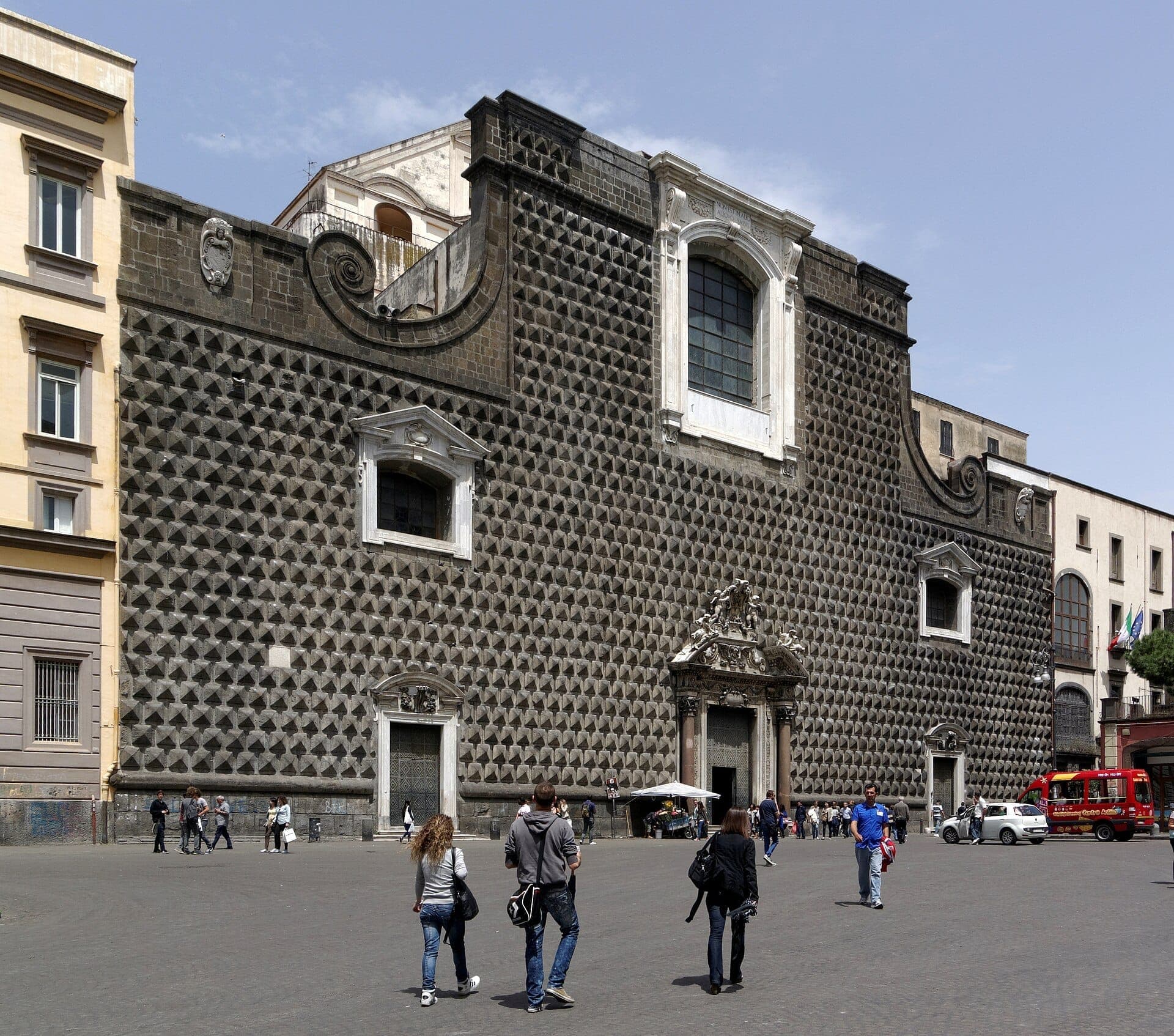 Chiesa del Gesù Nuovo em Nápoles com sua distintiva fachada de pedra em ponta de diamante, emoldurada por edifícios da cidade e visitantes caminhando nas proximidades em um dia ensolarado.