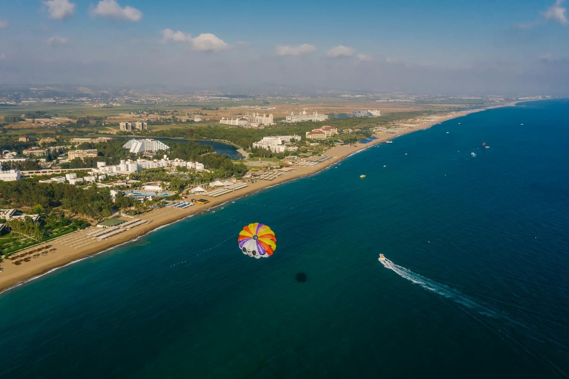Luftaufnahme eines mediterranen Küstenresorts in Antalya mit Hotels, Strand und einem Parasail über dem blauen Meer.