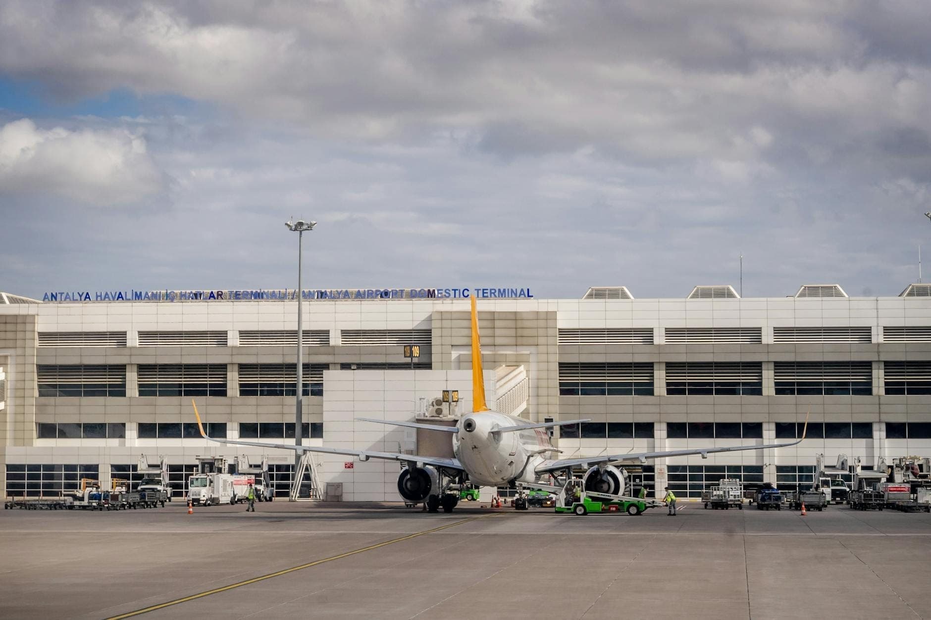 Inlandsterminal des Flughafens Antalya mit einem Passagierflugzeug auf dem Rollfeld vor dem Gebäude unter einem bewölkten Himmel.