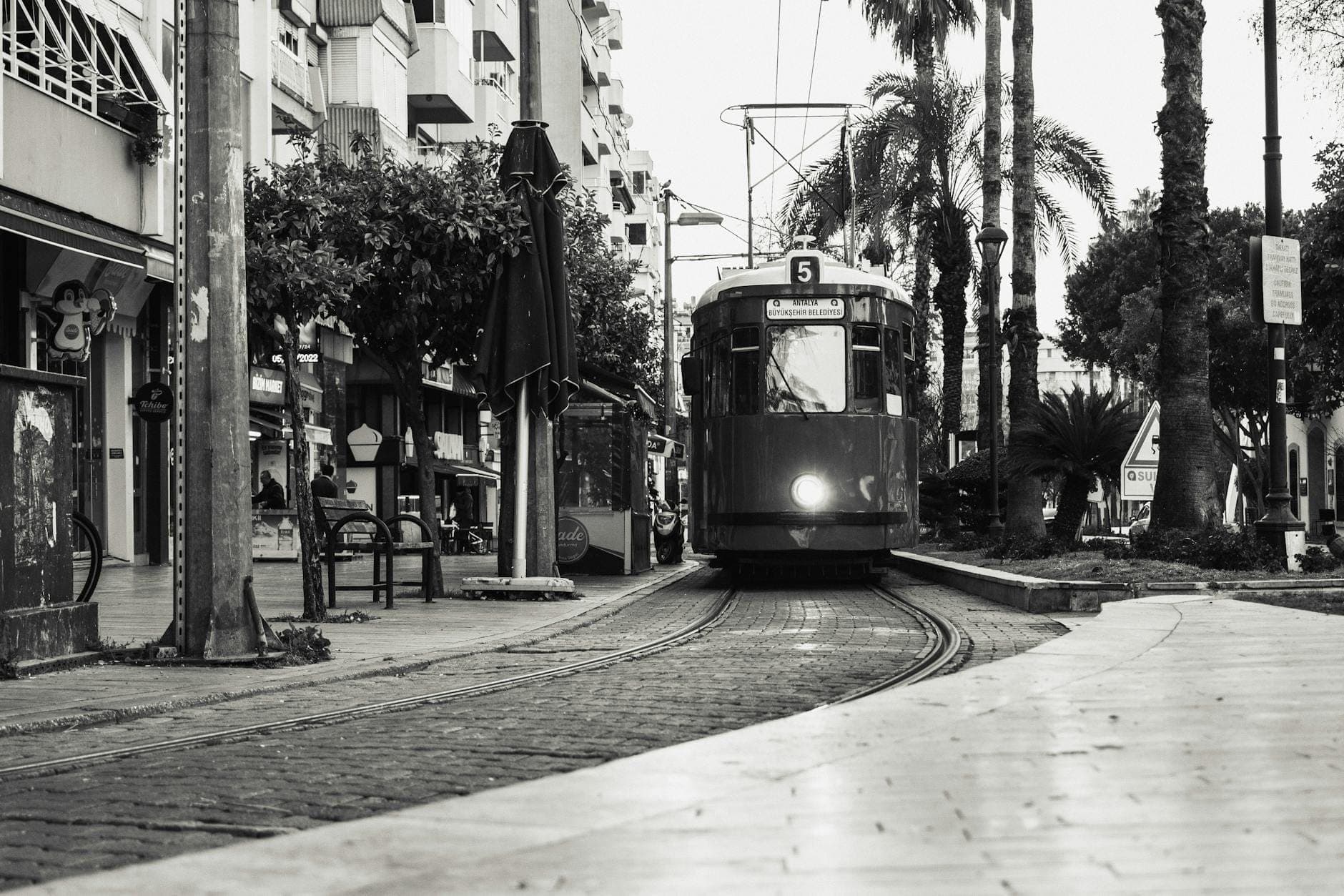Schwarz-Weiß-Foto einer heranfahrenden Straßenbahn im Zentrum von Antalya, mit Palmen, Gebäuden und Menschen auf der Straße.