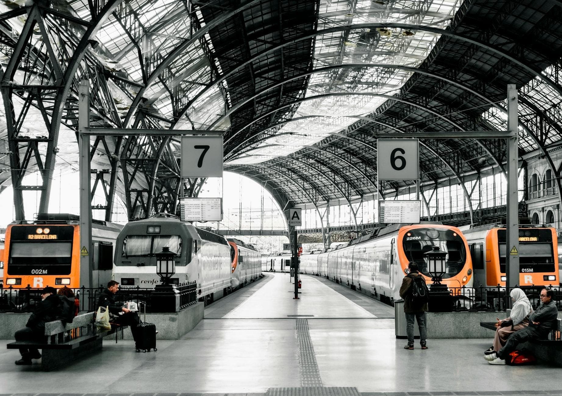 Pemandangan luas interior stasiun kereta Barcelona dengan beberapa kereta regional, langit-langit kaca besar, penumpang yang menunggu dan berjalan.