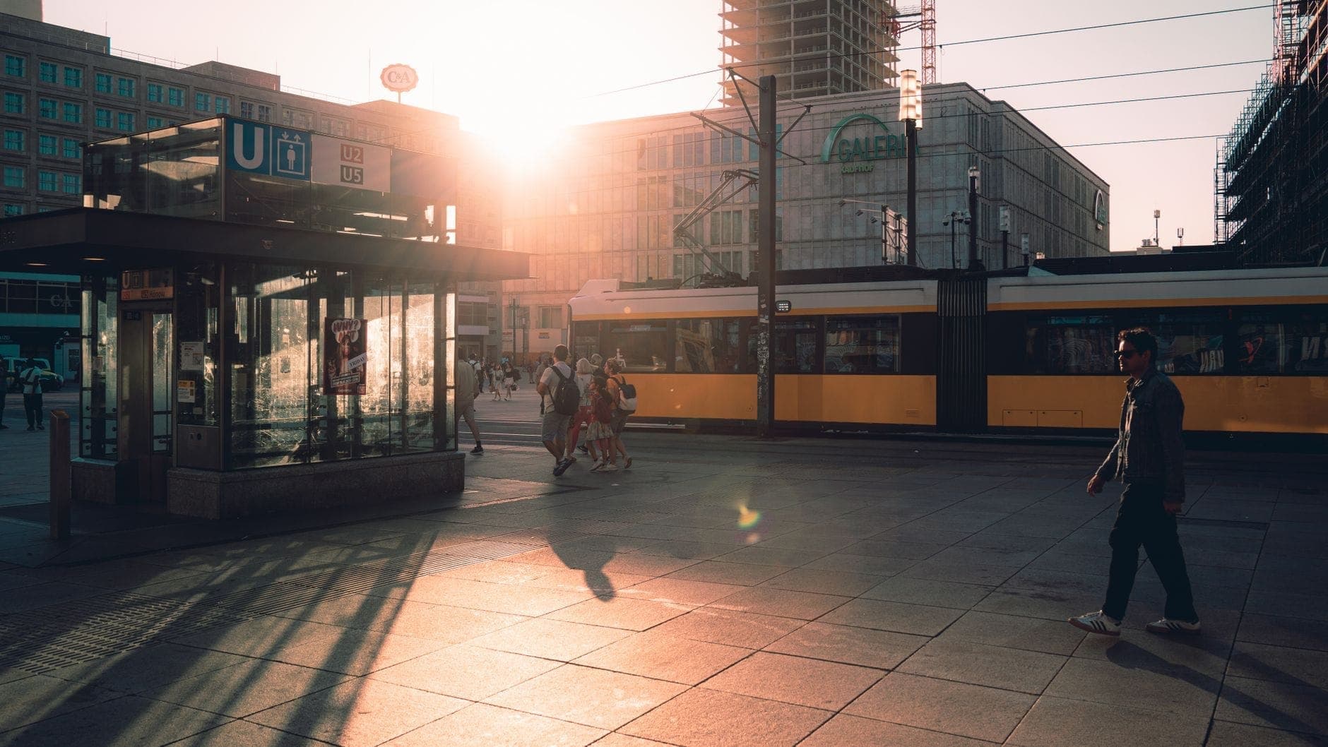 ภาพถนนในเบอร์ลินยามพระอาทิตย์ตกดิน พร้อมรถรางสีเหลือง ทางเข้า U-Bahn ผู้คนเดินอยู่ และอาคารในเมืองเป็นฉากหลัง