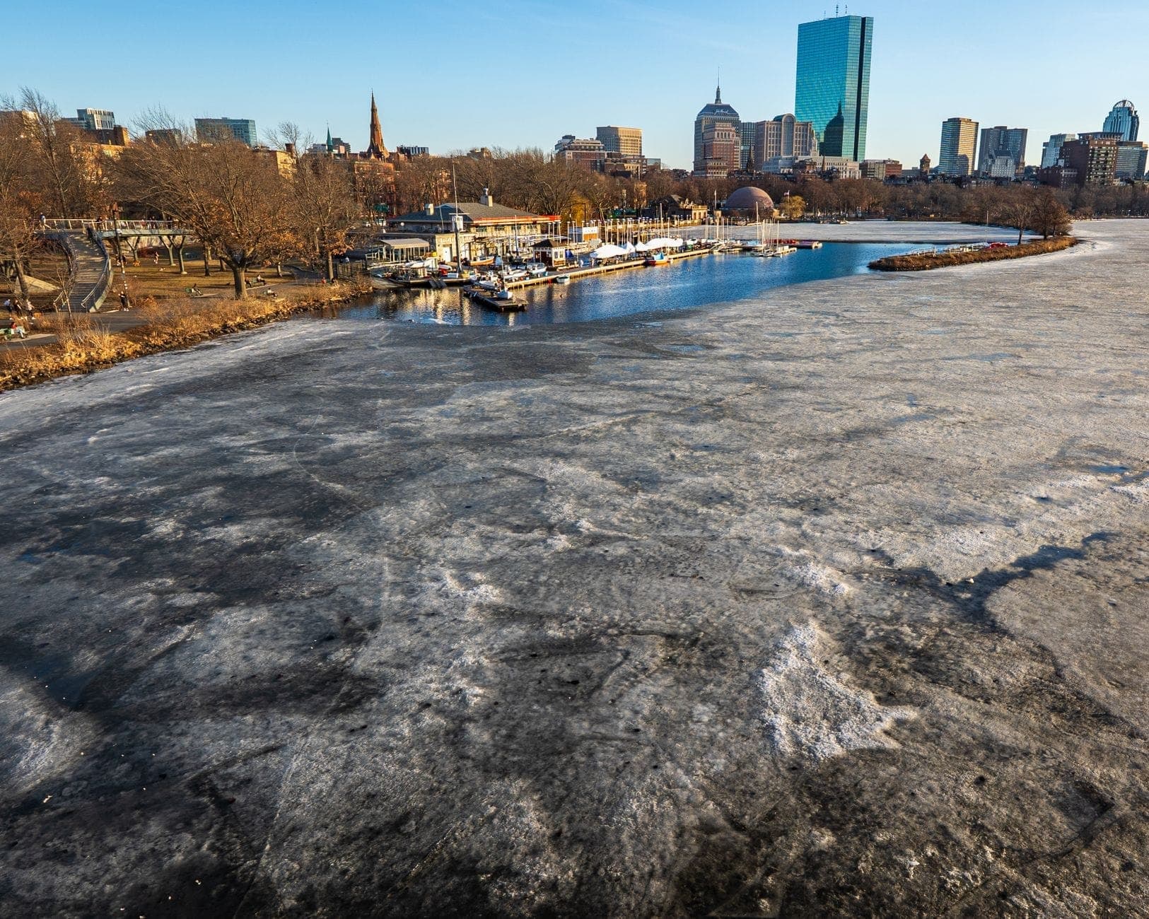 Charles River gelé avec des quais et la ligne d'horizon de Boston en arrière-plan, montrant des parties de la piste cyclable en bord de rivière.