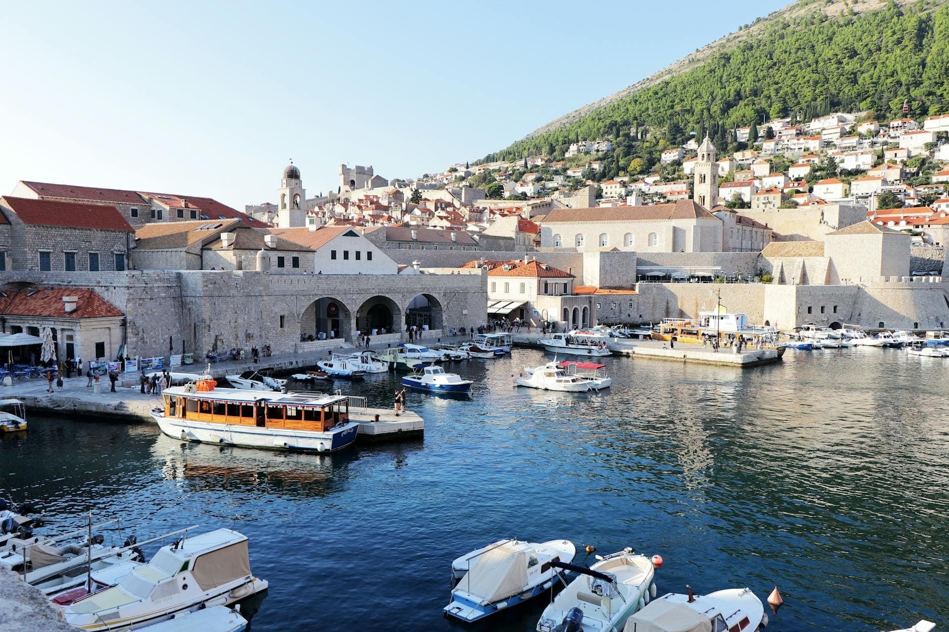 Boote und Fähren am alten Hafen von Dubrovnik mit Stadtmauern und historischen Gebäuden im Hintergrund im Sonnenlicht.